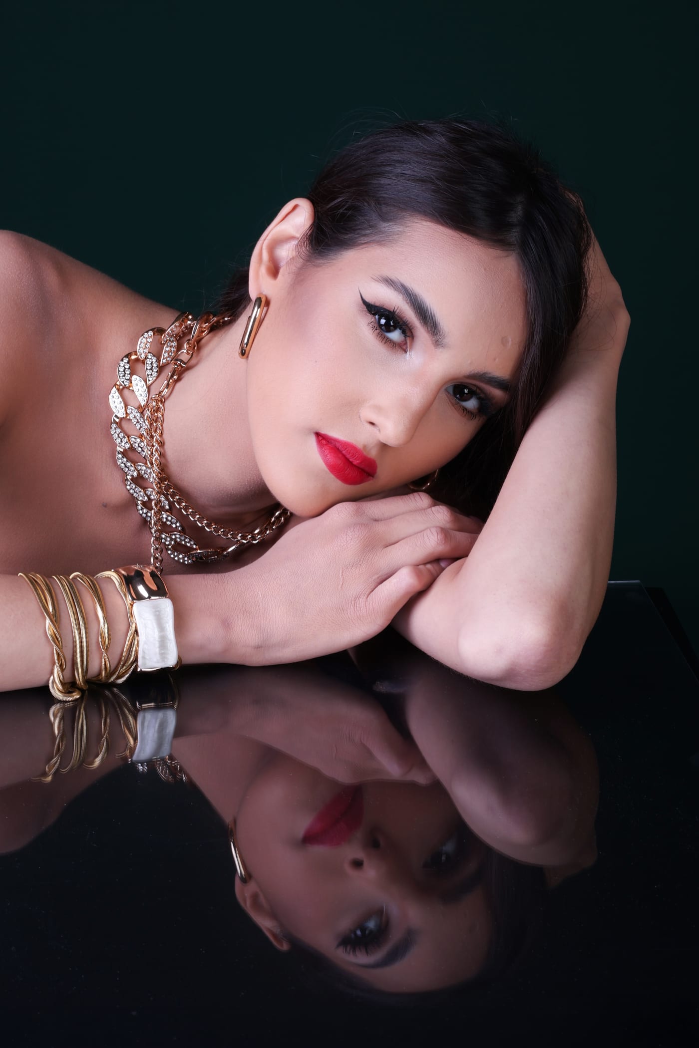 Beauty portrait in gold jewelry, head resting on the arm, mirrored on black glass