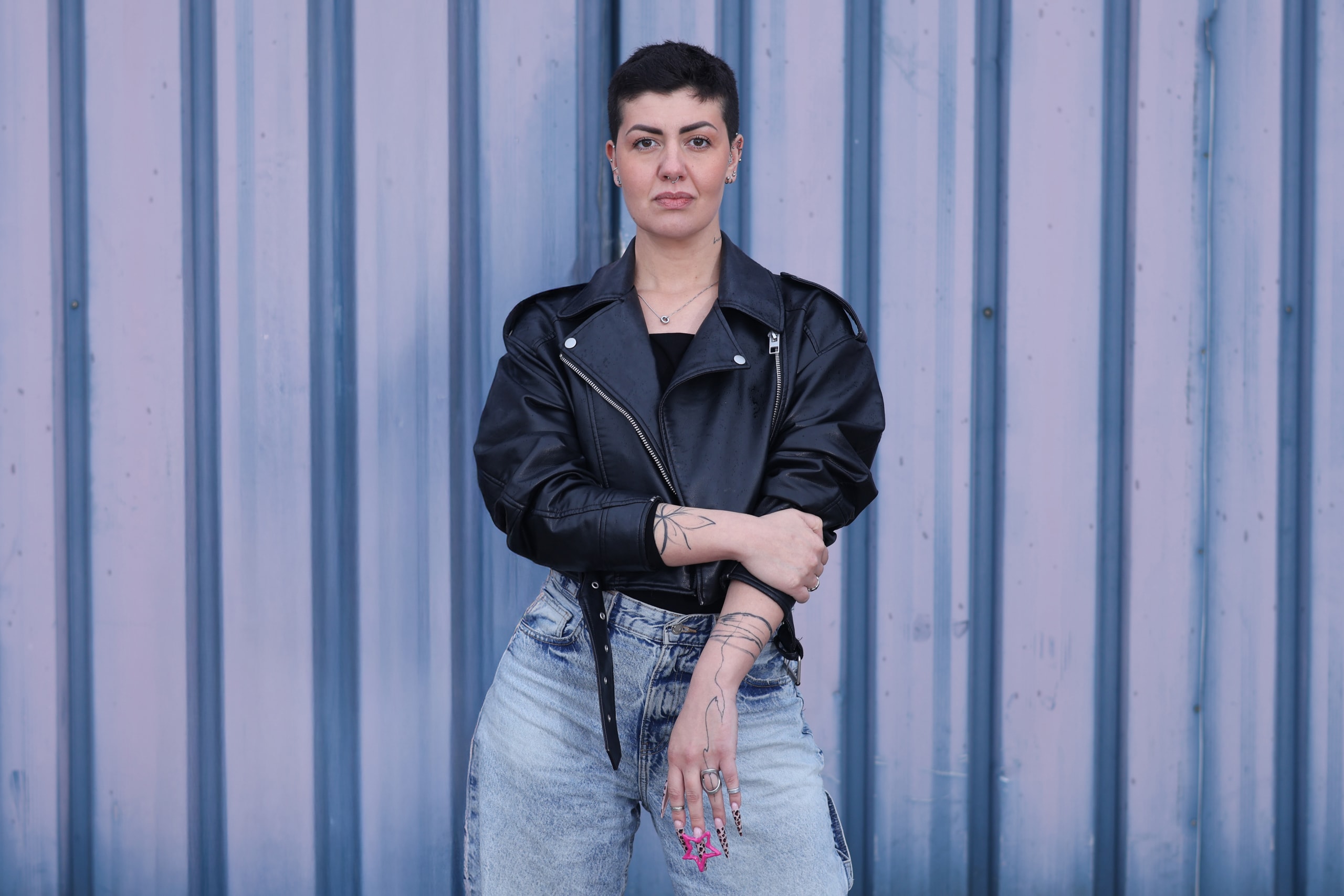 Half-length against the ribbed wall, arms crossed low, leather jacket, direct gaze