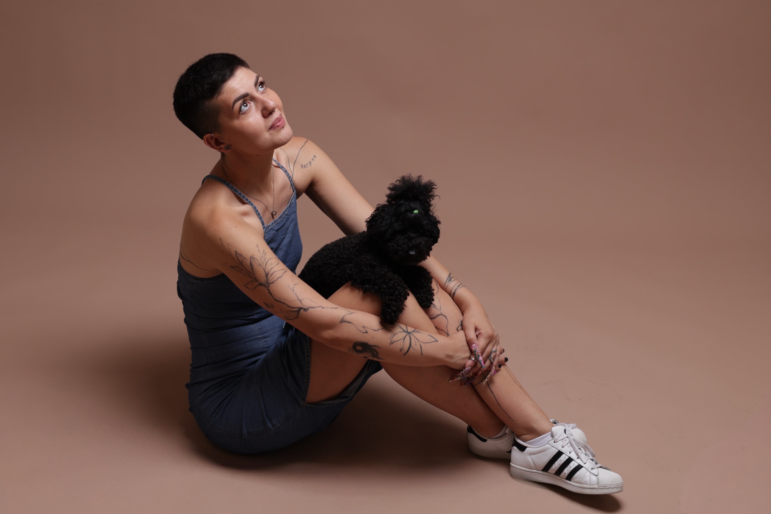 Seated on the floor against warm tan, holding a small black dog, white sneakers