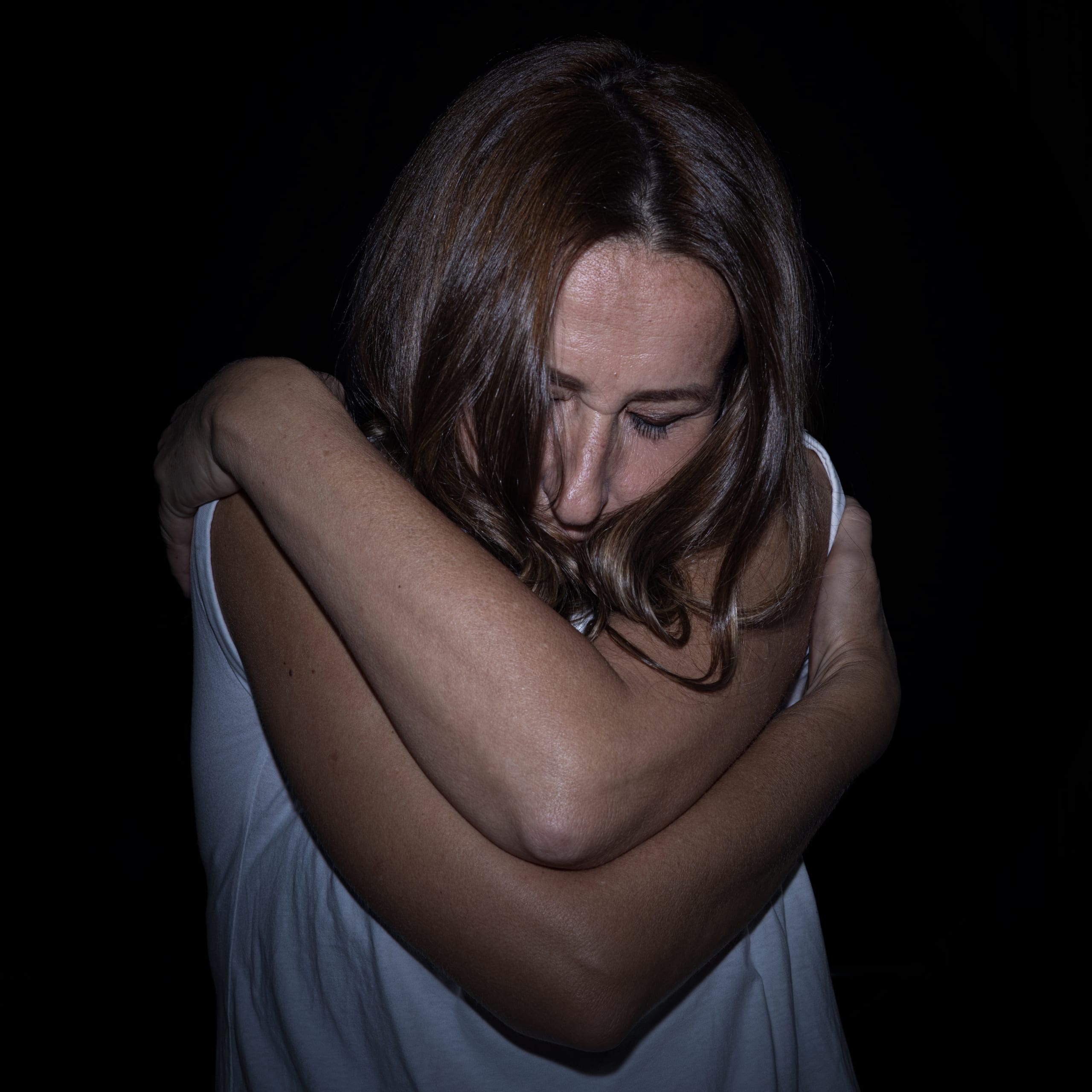 A woman in a white top holding herself, face turned down in shadow