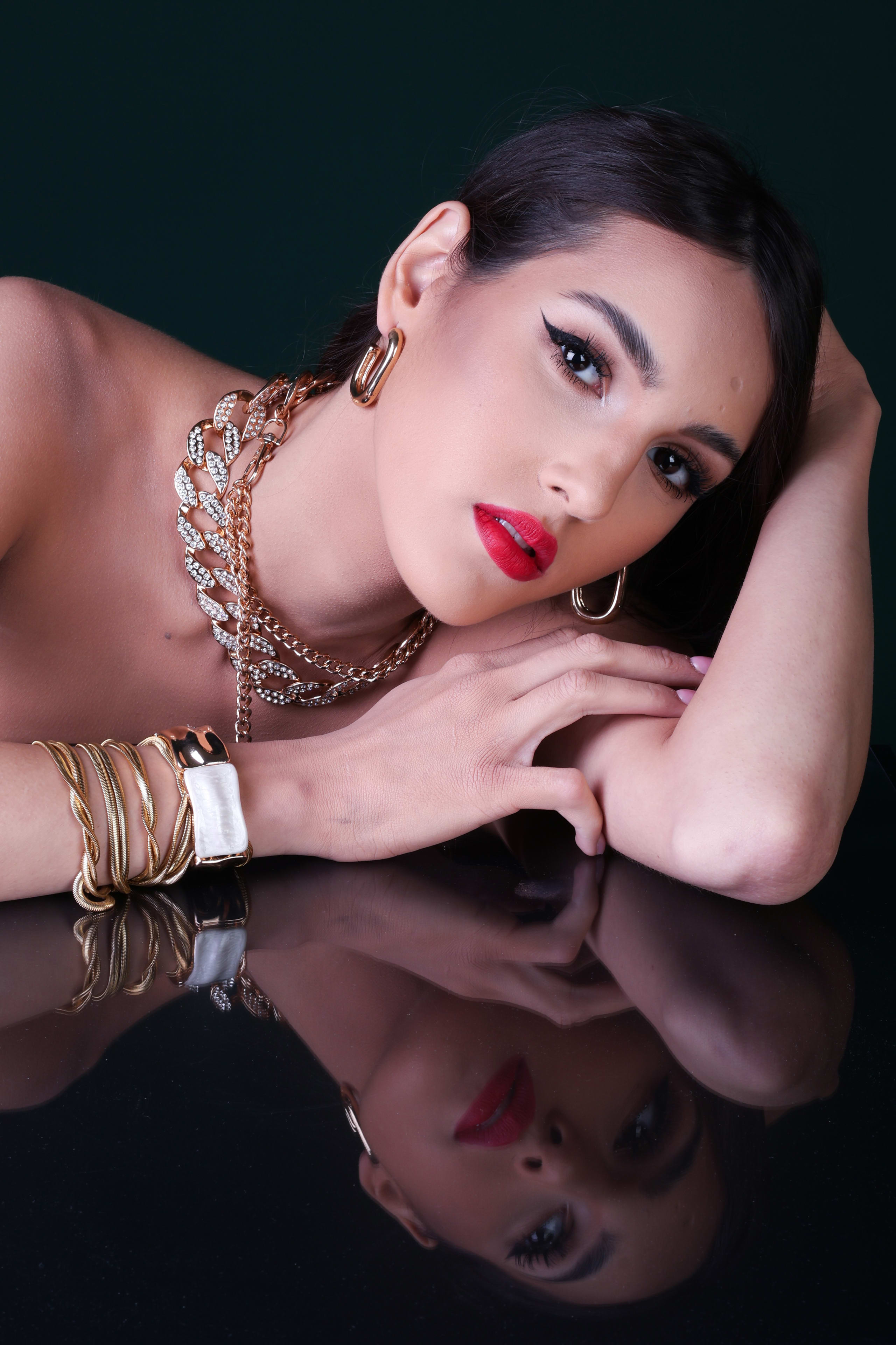 Head resting on her arm, red lip and direct gaze, gold chain necklaces and a gold cuff, reflected below