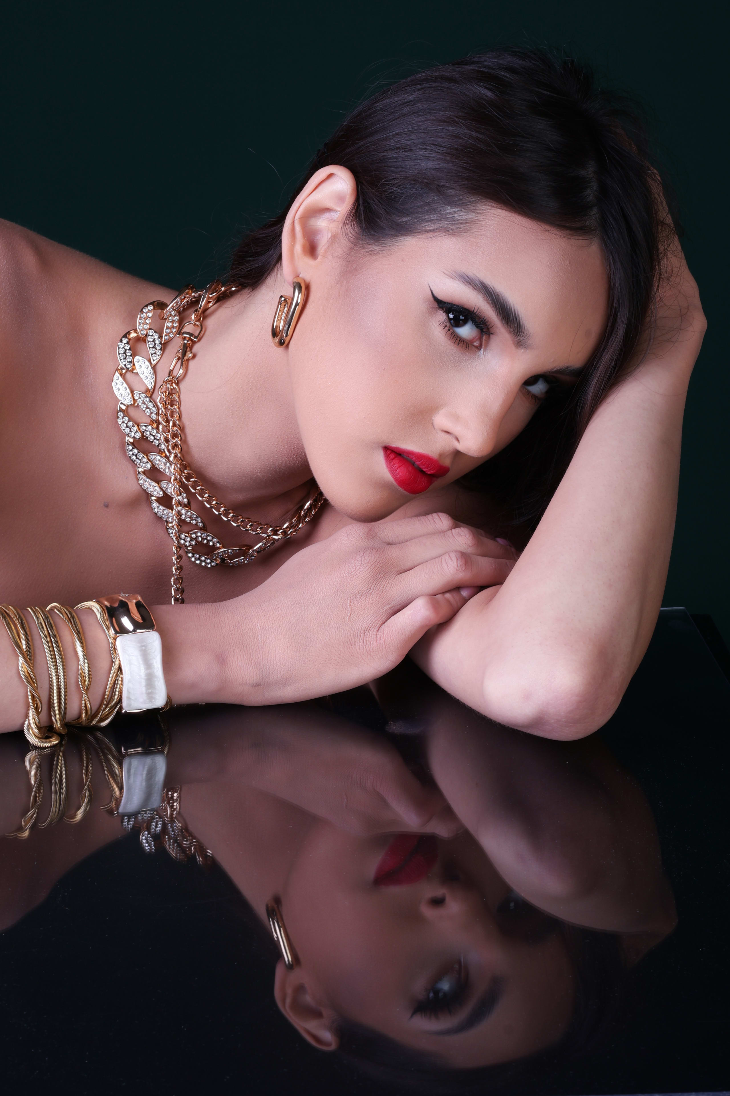 Chin lifted with a direct gaze, layered gold necklaces and hoop earrings, reflection on the black mirror