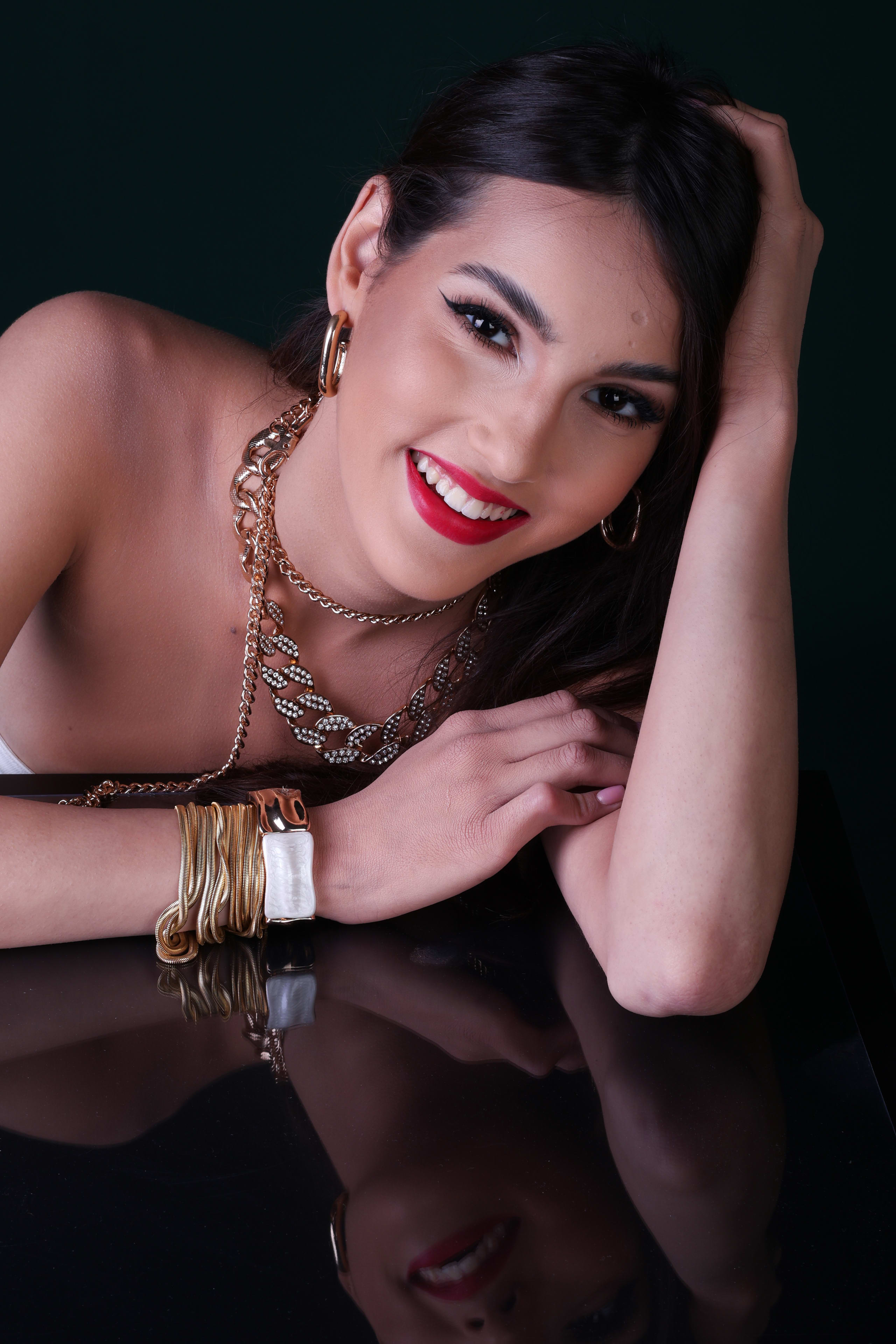 Warm smile with a hand in her hair, gold chains and a white-and-gold cuff, mirrored on black glass