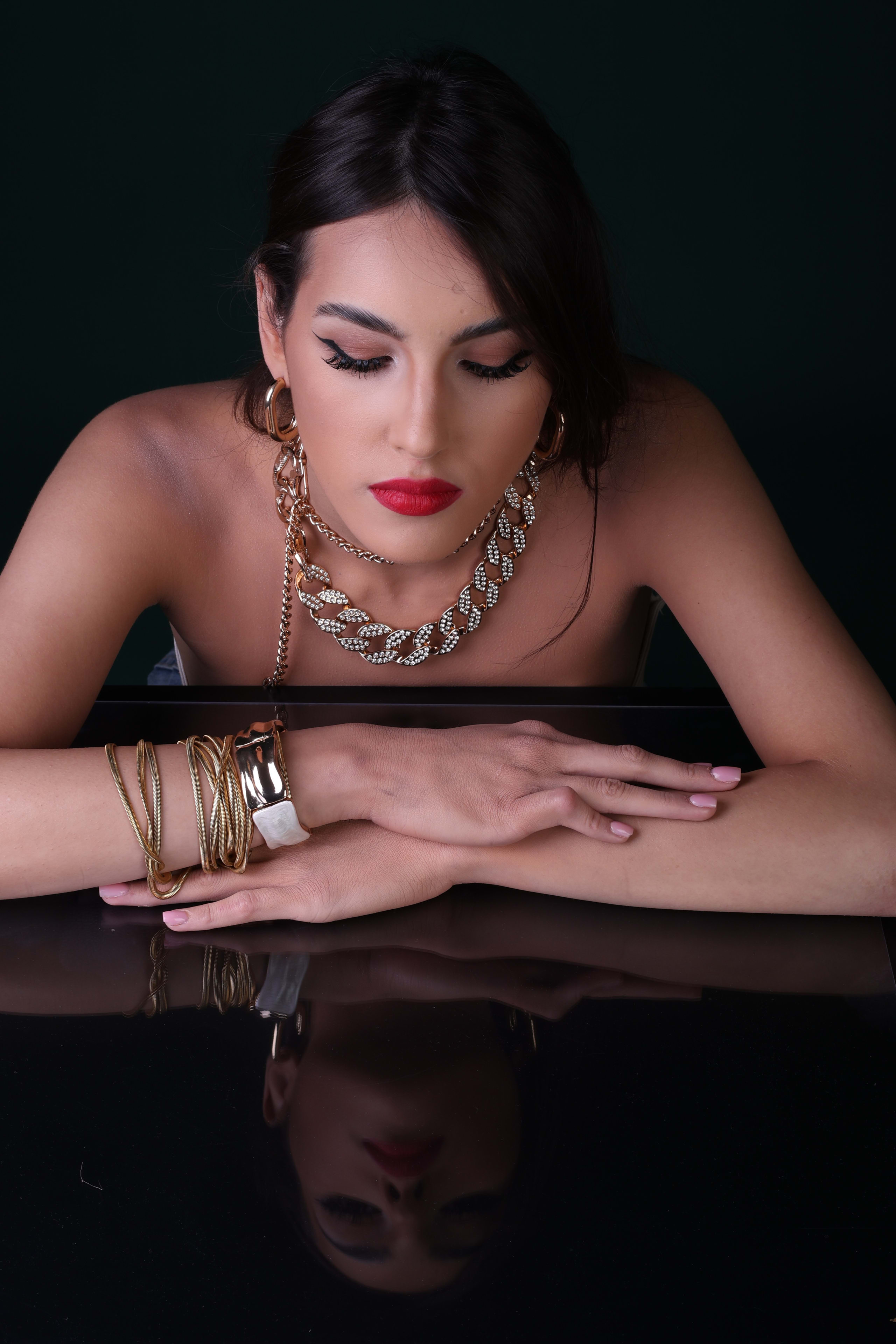 Eyes lowered, resting on folded hands, gold chain-link necklace, a calm reflection below