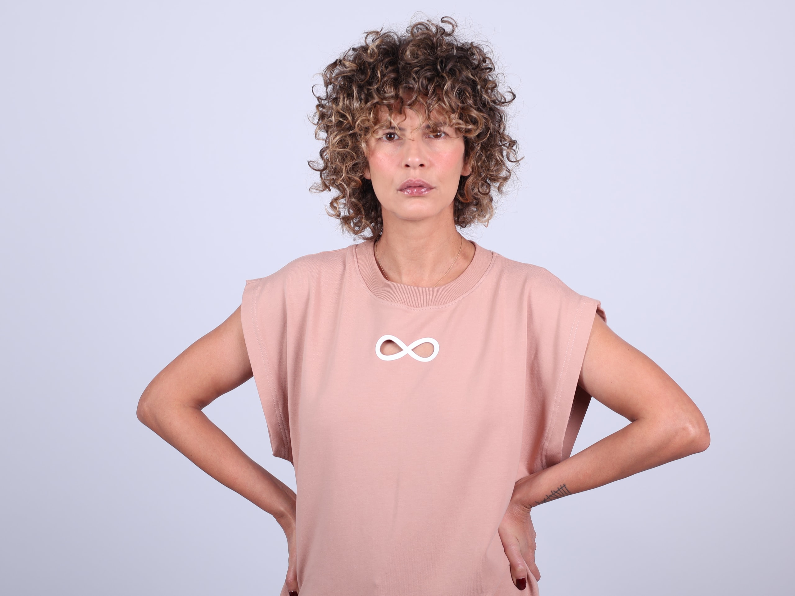Standing with hands on hips in the dusty-pink top, curly hair, a defiant gaze