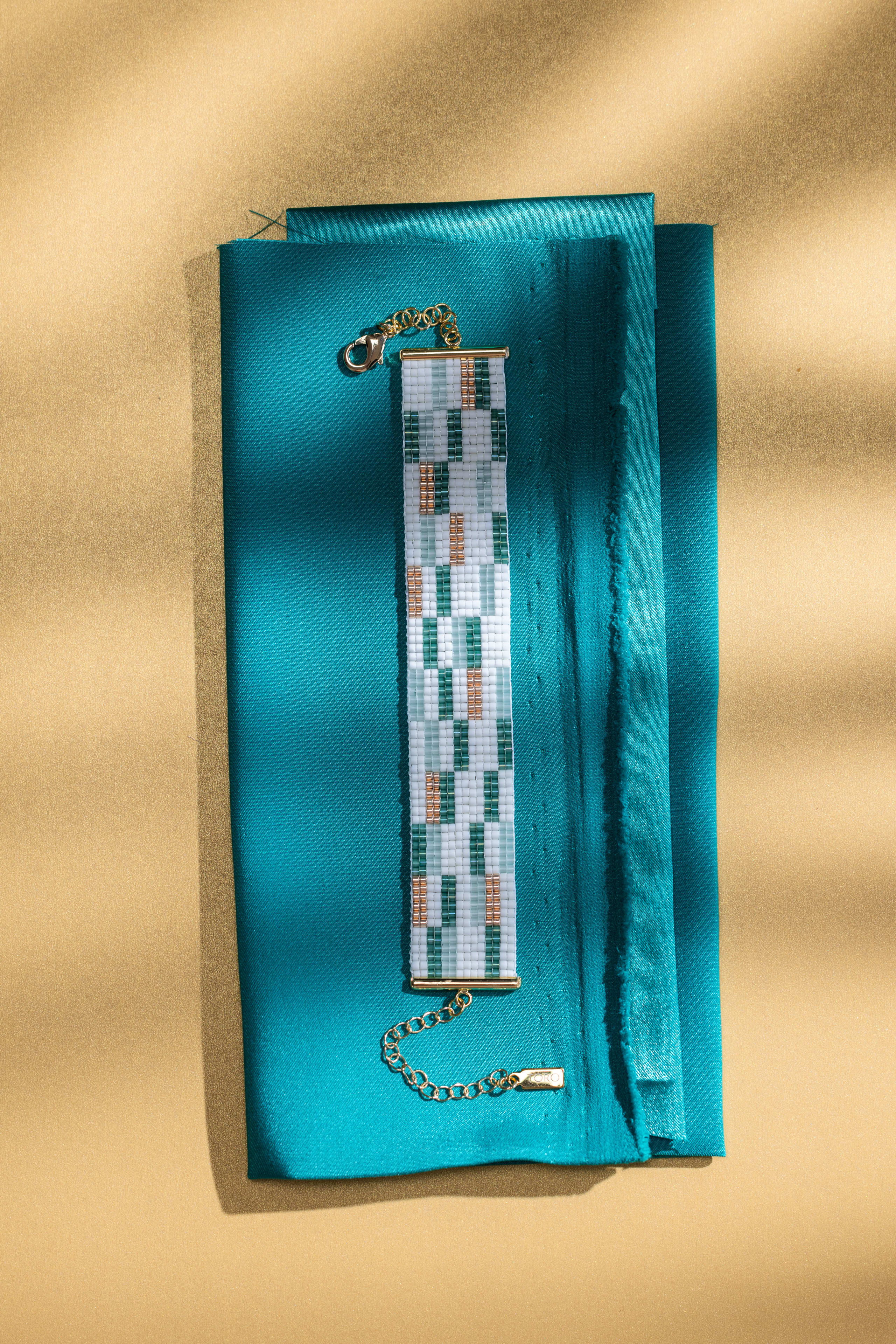 A green and white checkered cuff on folded teal silk, diagonal shadows falling across the gold ground behind