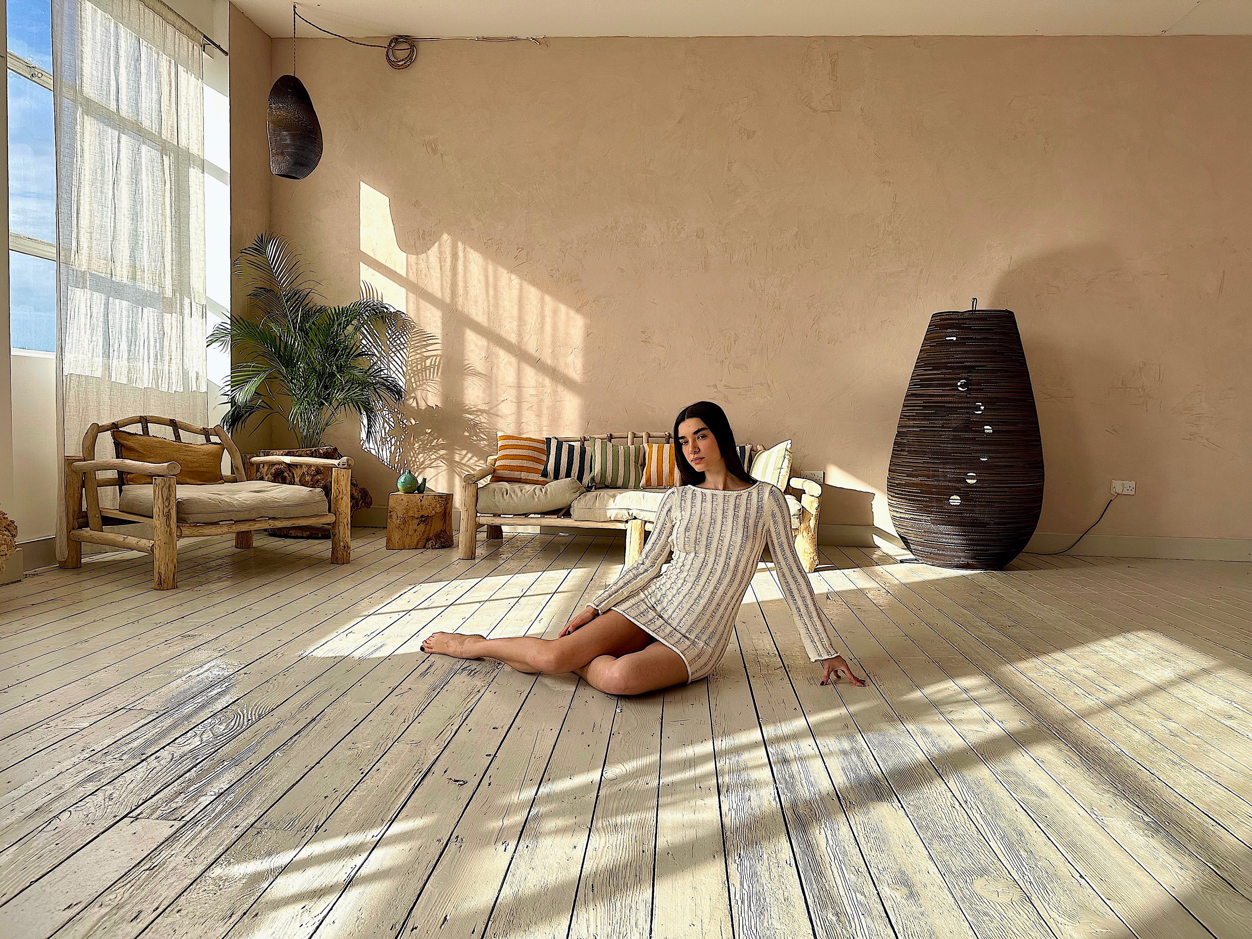 A woman poses casually in a wooden chair by a large window, with natural light illuminating the room's organic decor.
