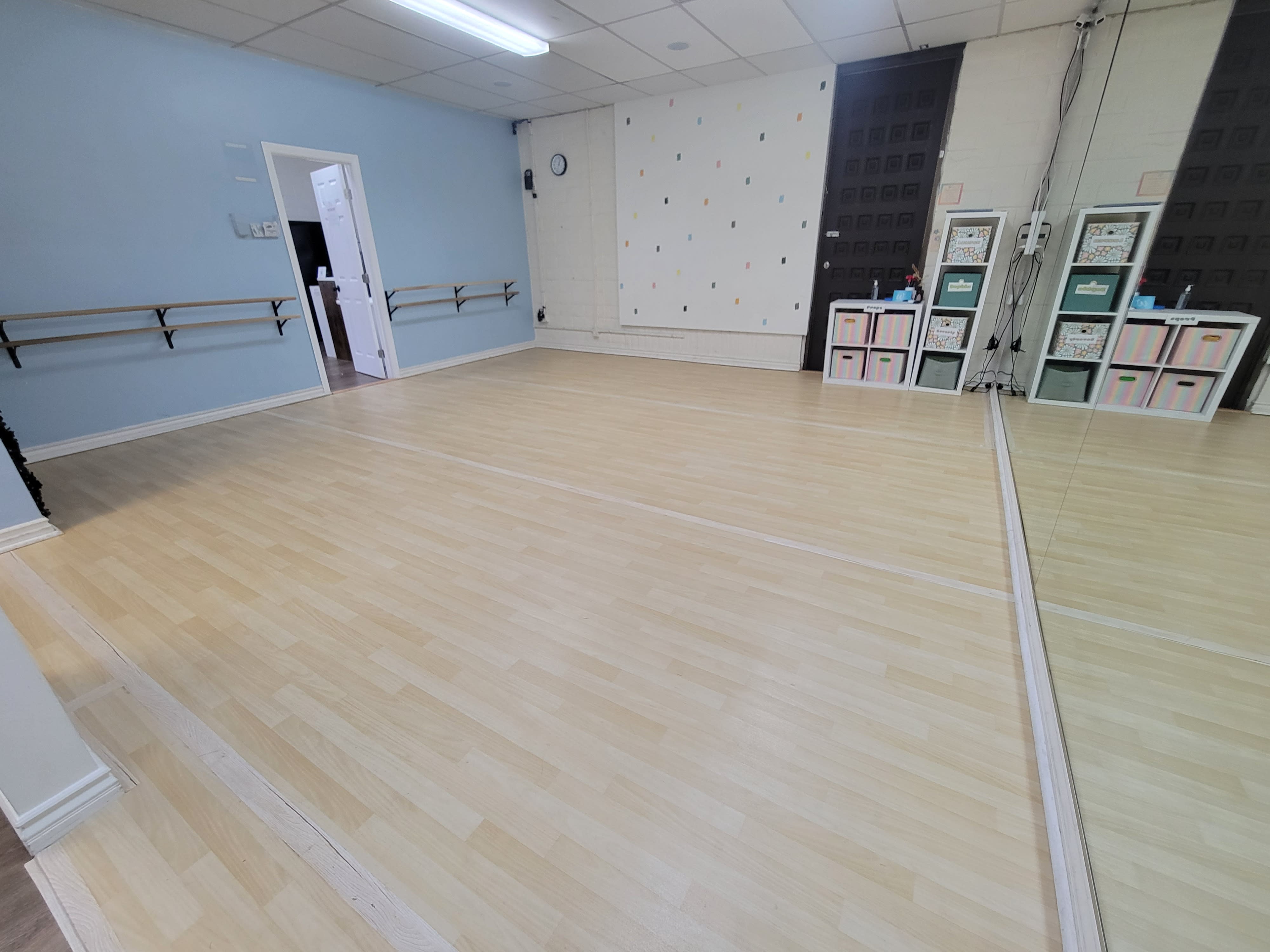 A dancer sits on the floor in a studio, reflecting in a mirror while surrounded by a laptop, water bottle, and a fan.