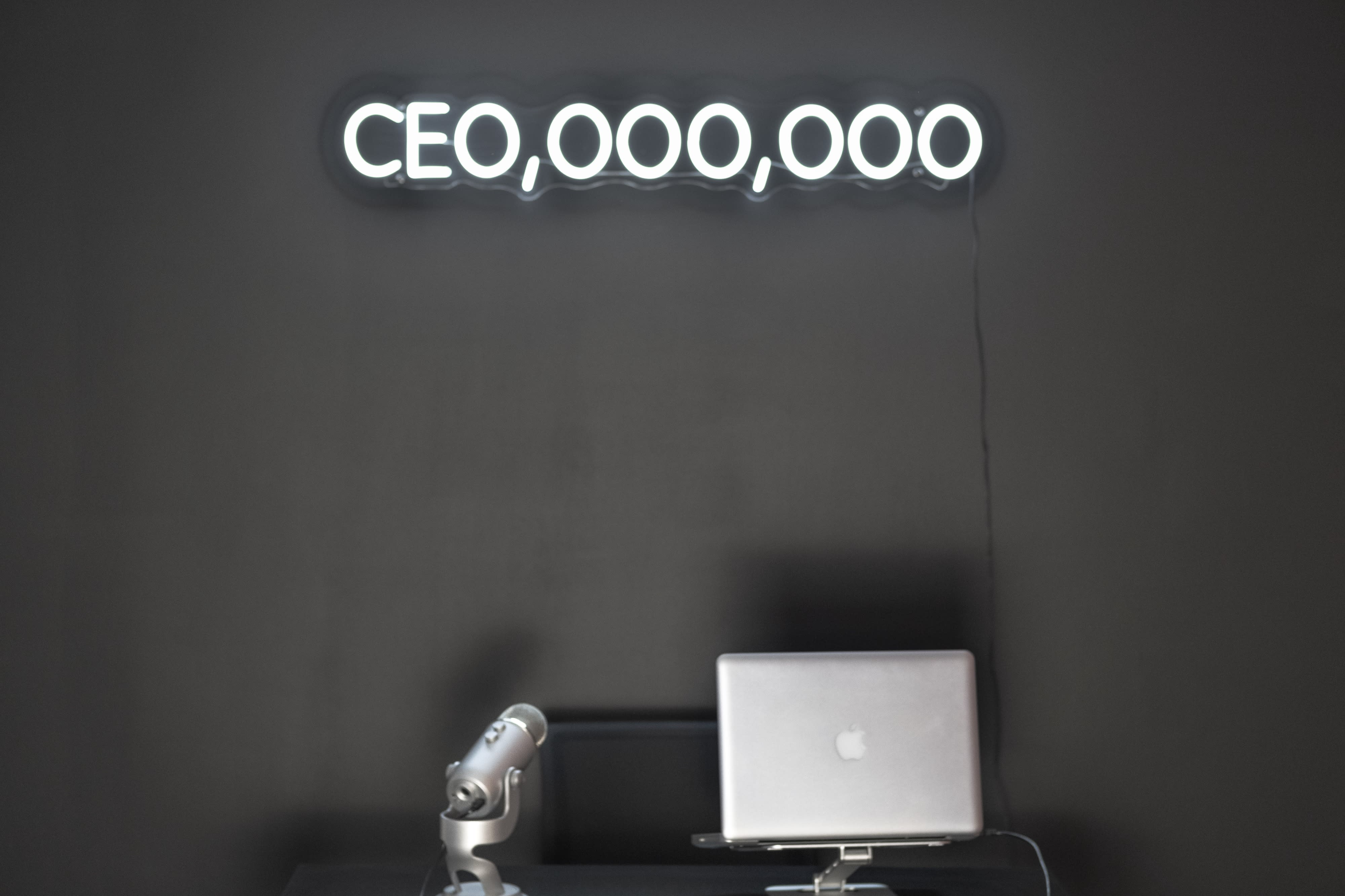 A person poses enthusiastically under a neon sign that reads "CEO,000,000," with a microphone on the table in front of them, indicating a podcast recording session.