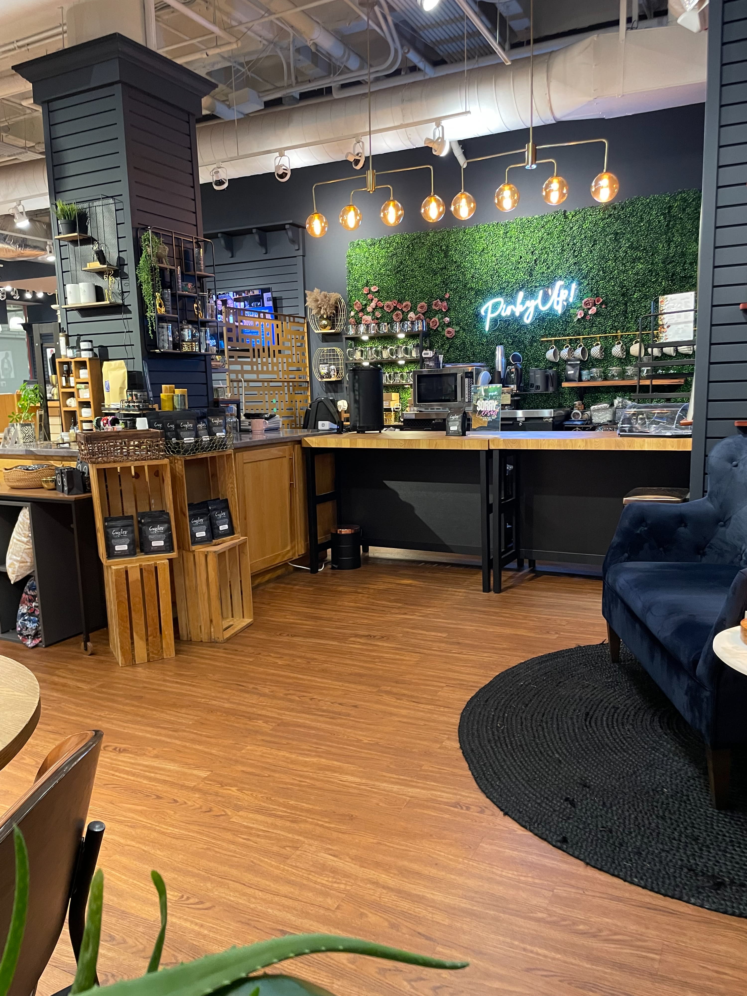 A display of travel-themed pins and a package of coffee are set on a wooden counter, with tea brewing in the background and a plant nearby.
