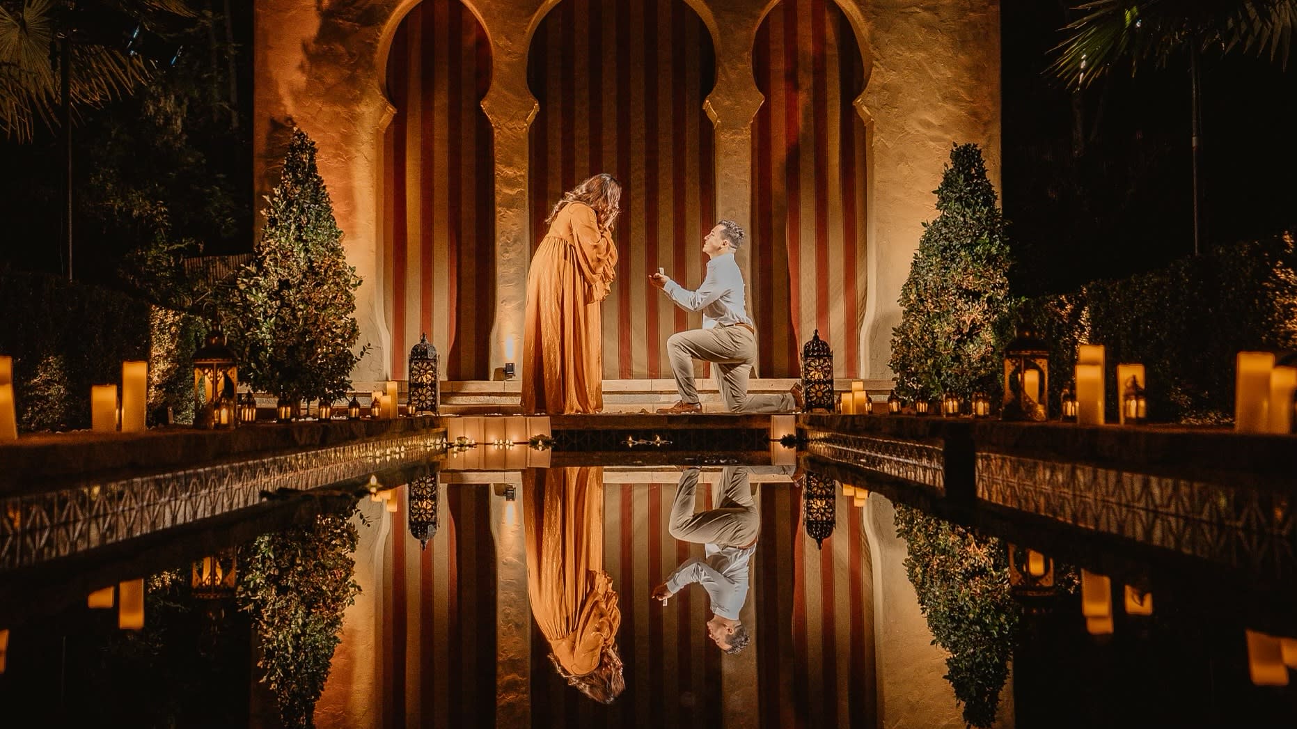 A man kneels in front of a woman, proposing marriage under a softly lit archway surrounded by candles and reflecting water.