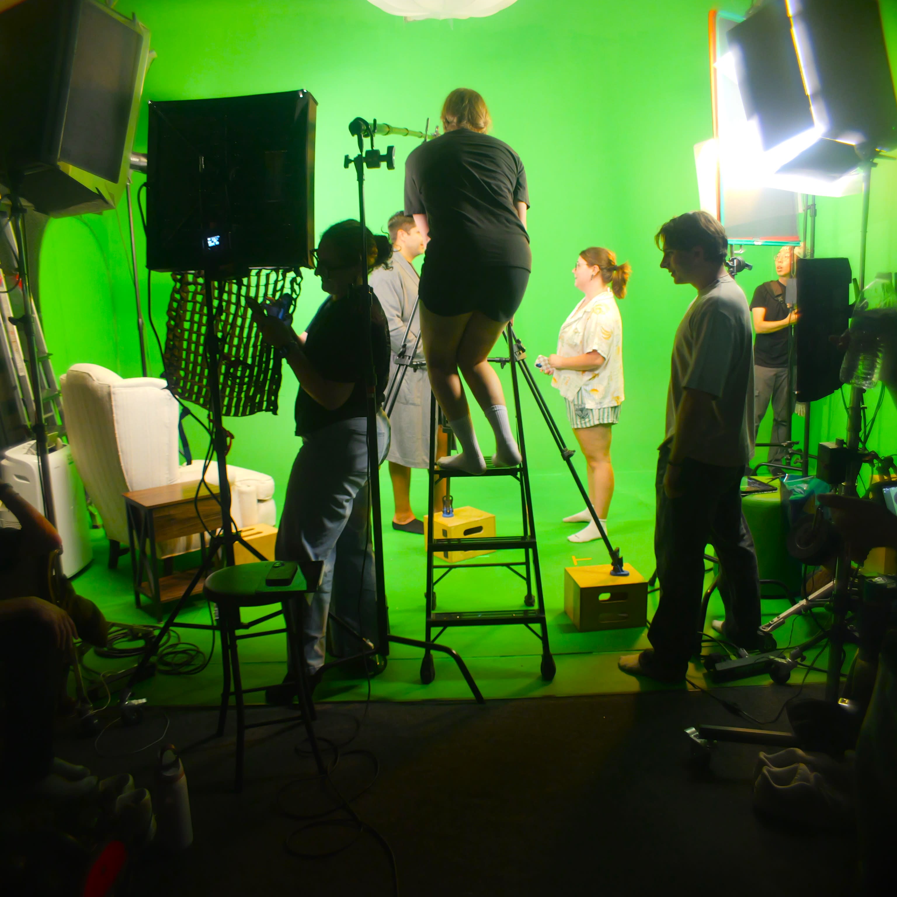 An elderly man sits in an armchair against a green screen, pointing while being filmed with a camera and microphone equipment set up around him.