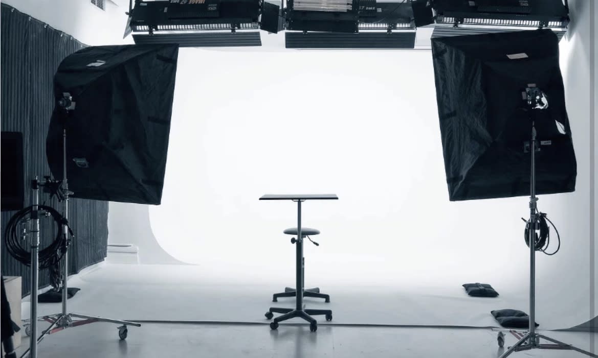 A production crew sets up cameras and monitors for a video shoot in a white studio while two individuals prepare for a scene.