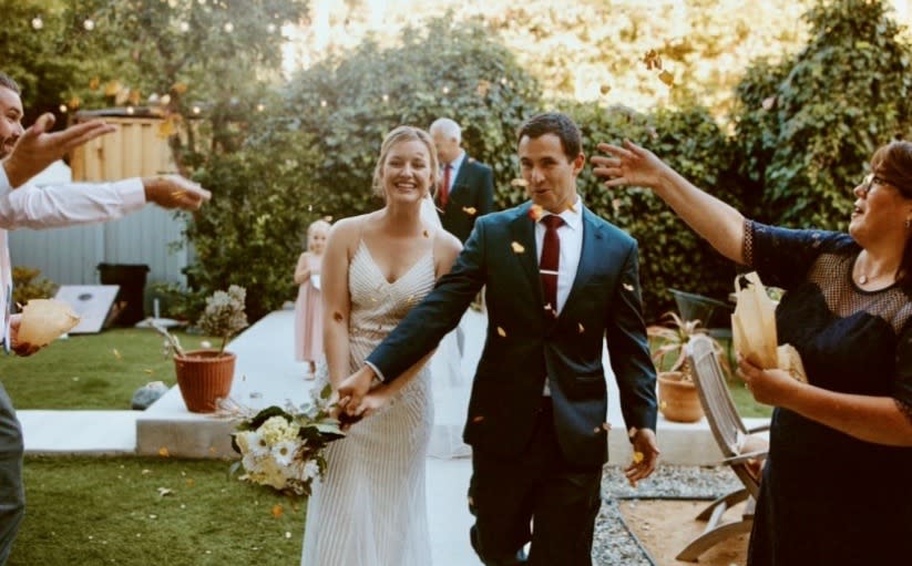 A groom twirls his bride in a white lace wedding dress outside a blue building, surrounded by desert plants.