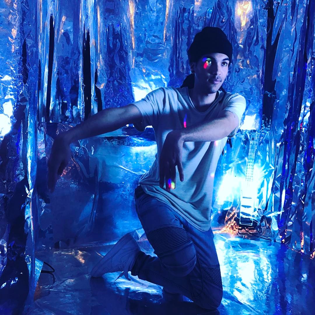 A film crew sets up a shoot in a reflective, metallic space with neon lights and cameras positioned on tripods.