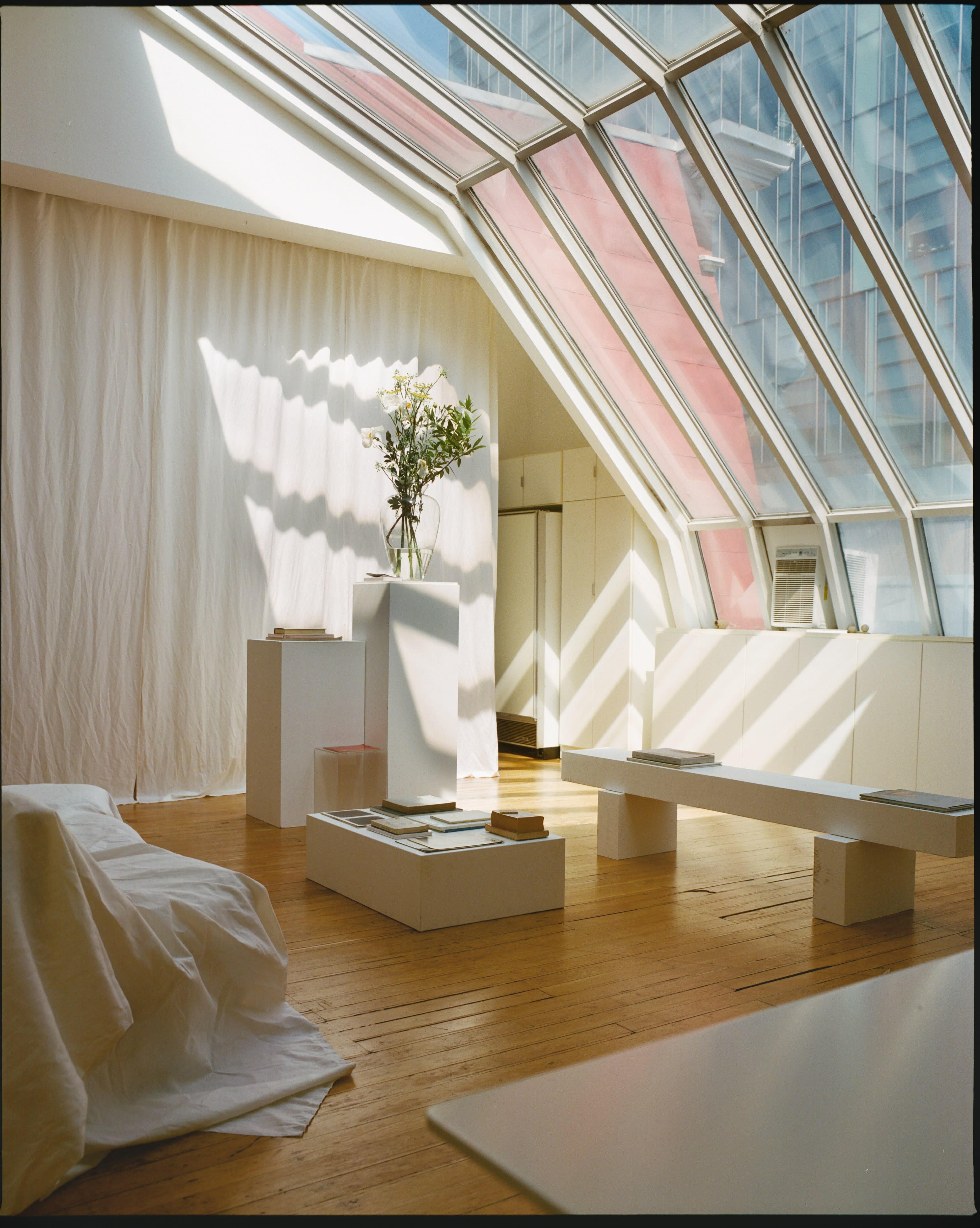 The scene features a bright, minimalist room with sheer fabric draping from the ceiling, a white sofa, and a simple wooden dining table surrounded by chairs.