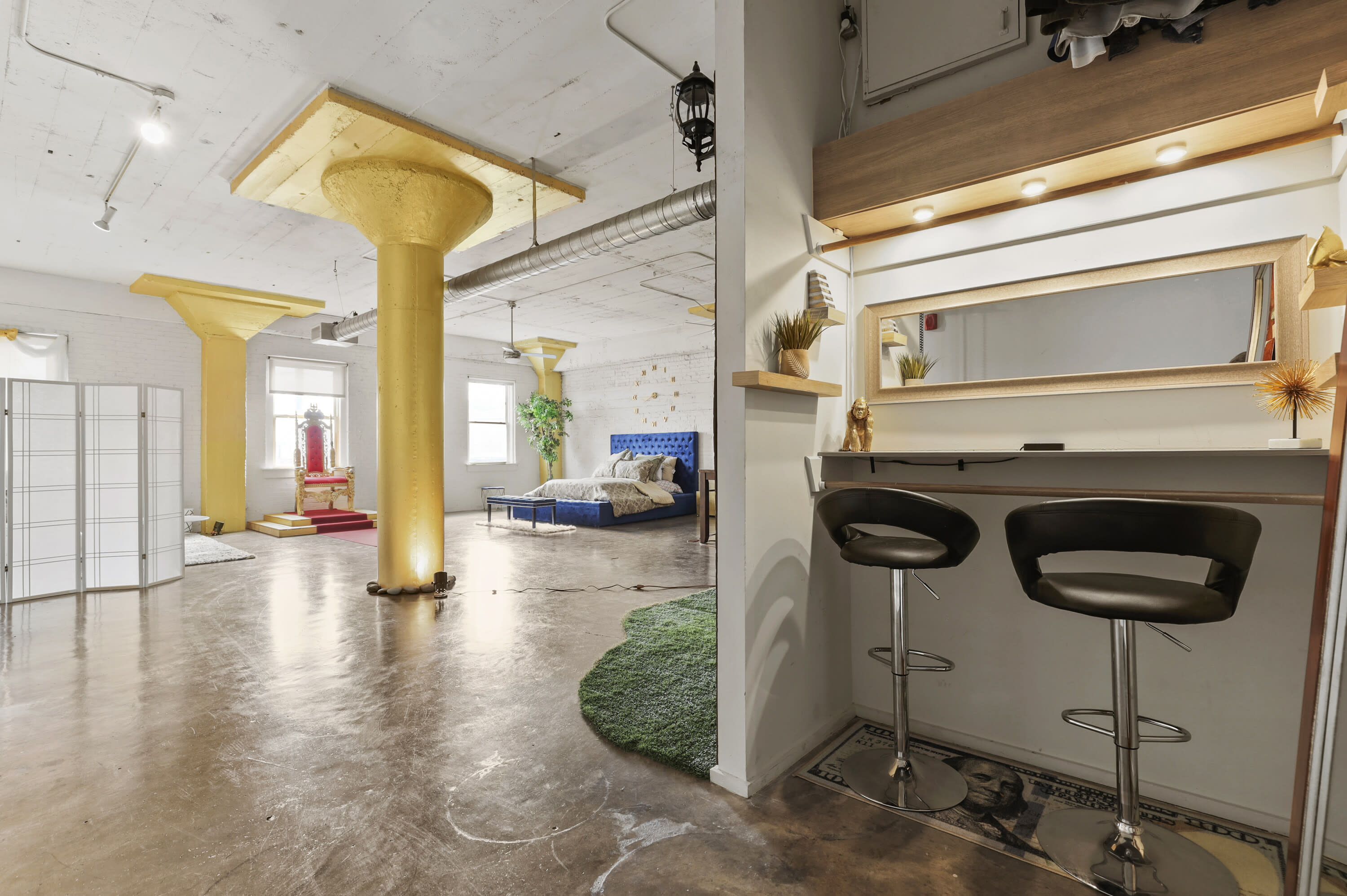 A photoshoot is taking place in a bright studio featuring a seating area and a bed setup, with lighting equipment positioned strategically around the space.