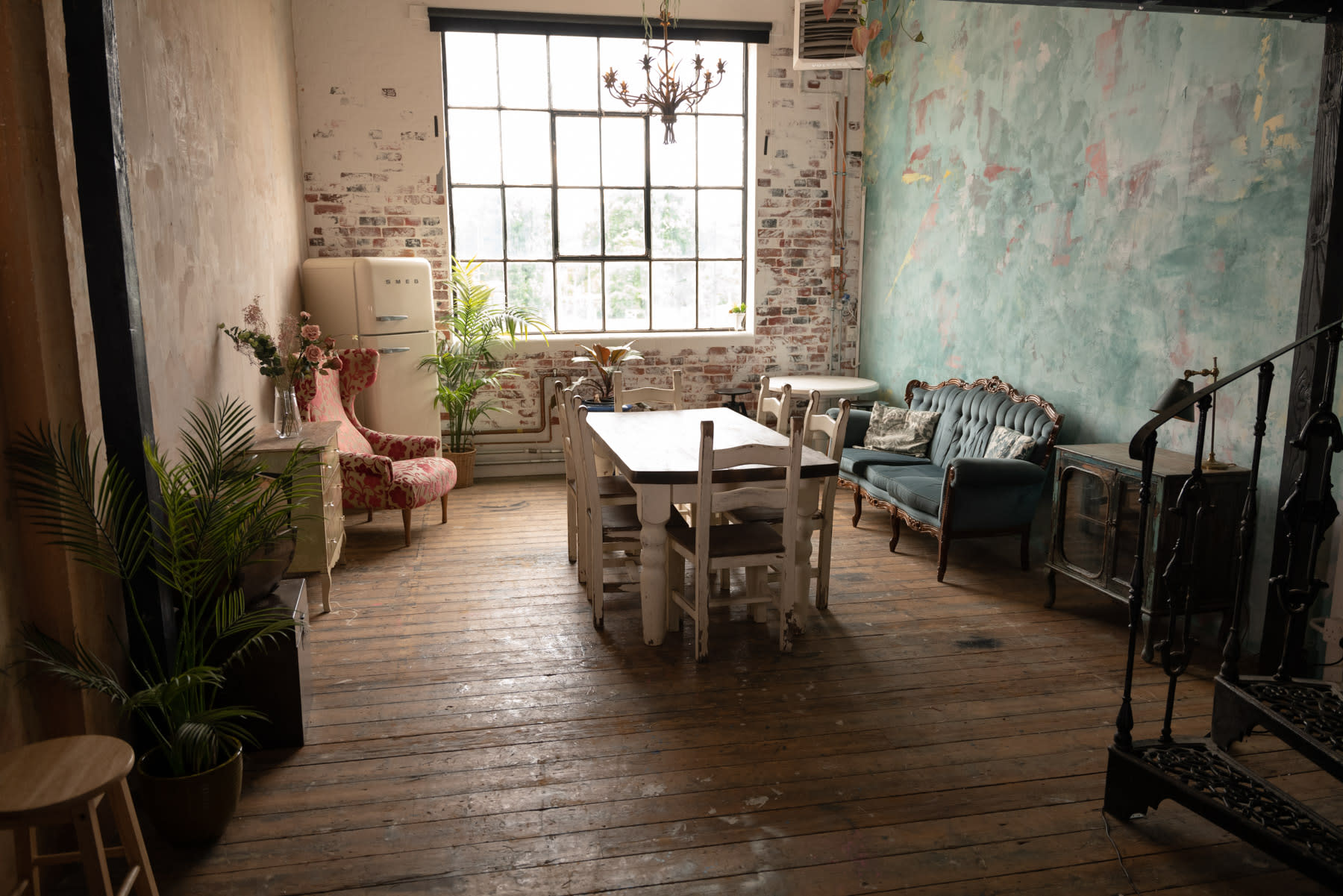 A film shoot takes place in an industrial-style room featuring a large table surrounded by vintage furniture, with natural light streaming through a large window.