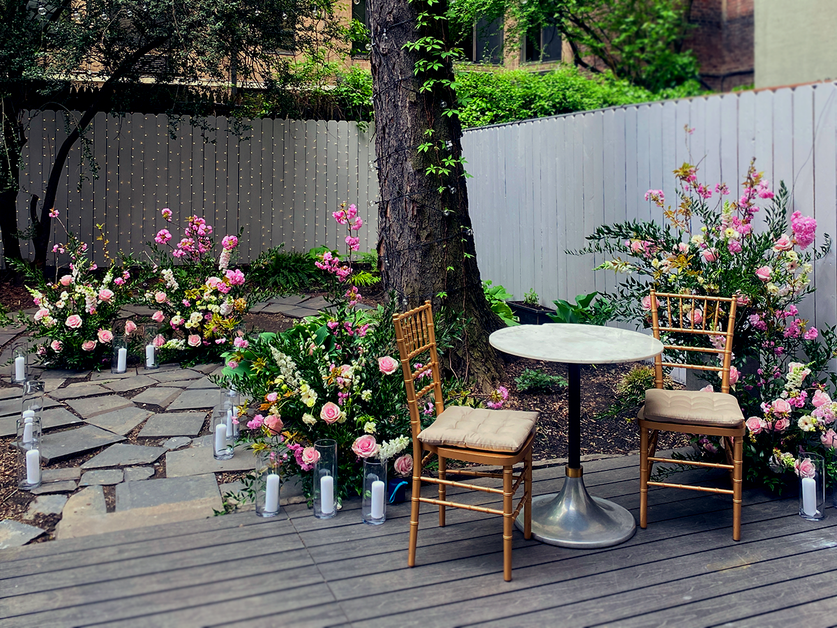 A group of five women poses together in a garden setting during a baby shower, wearing hats and festive attire.