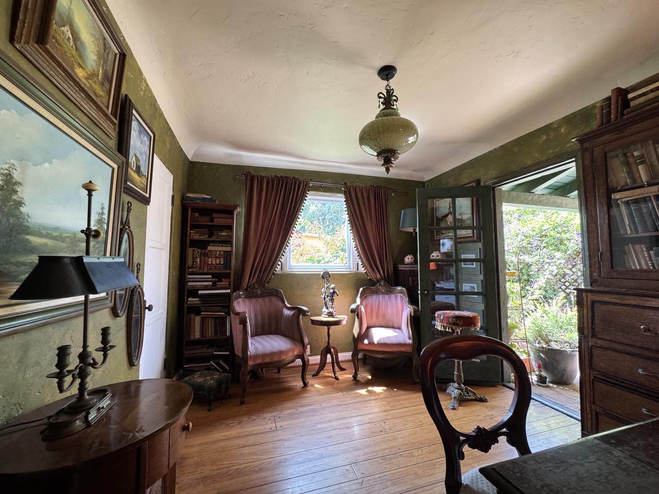 A person sits on a mantelpiece in a cozy room filled with tall bookshelves, surrounded by plants and natural light.