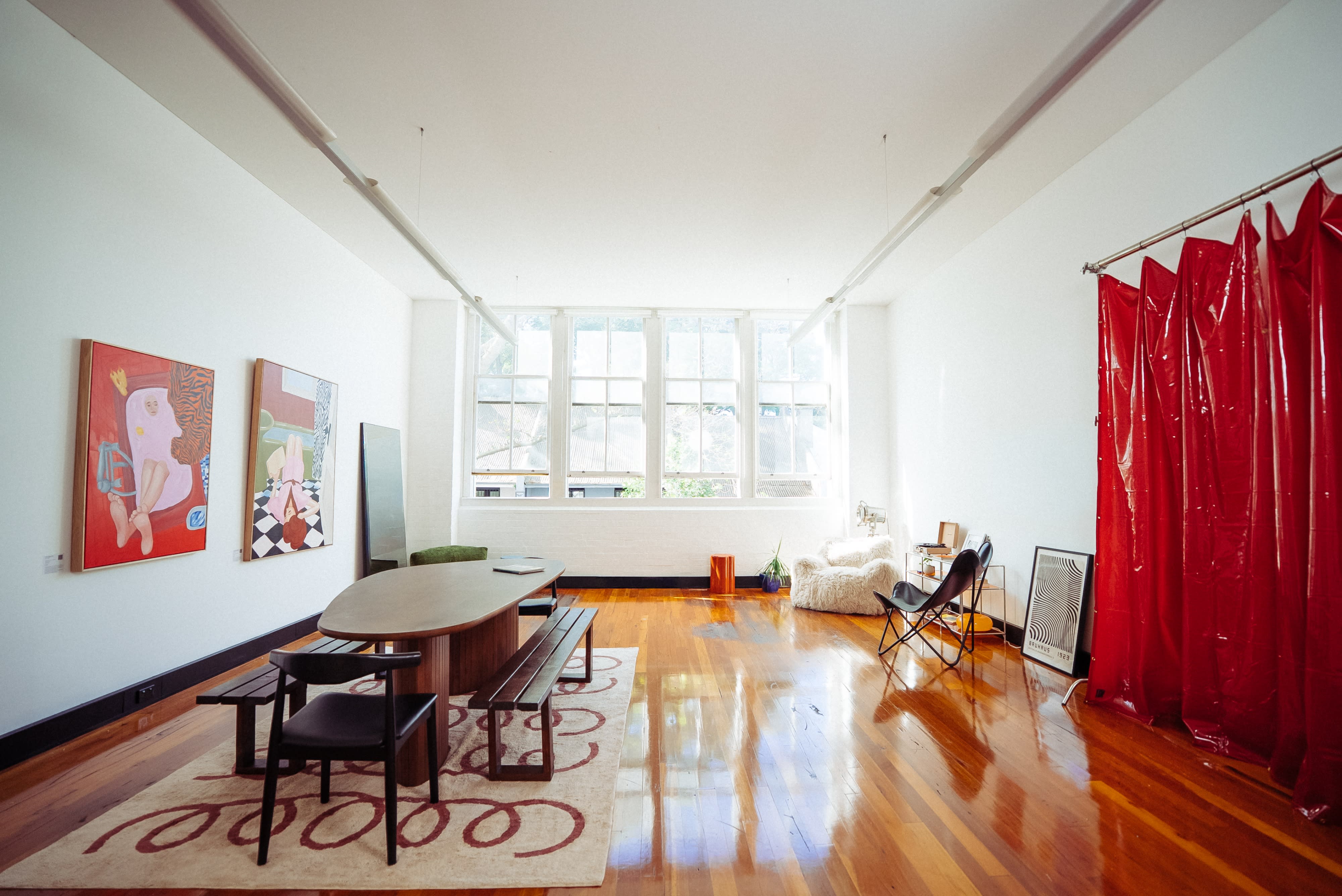 A spacious room features a large table with benches, surrounded by large windows allowing natural light, and a cozy seating area in the corner.