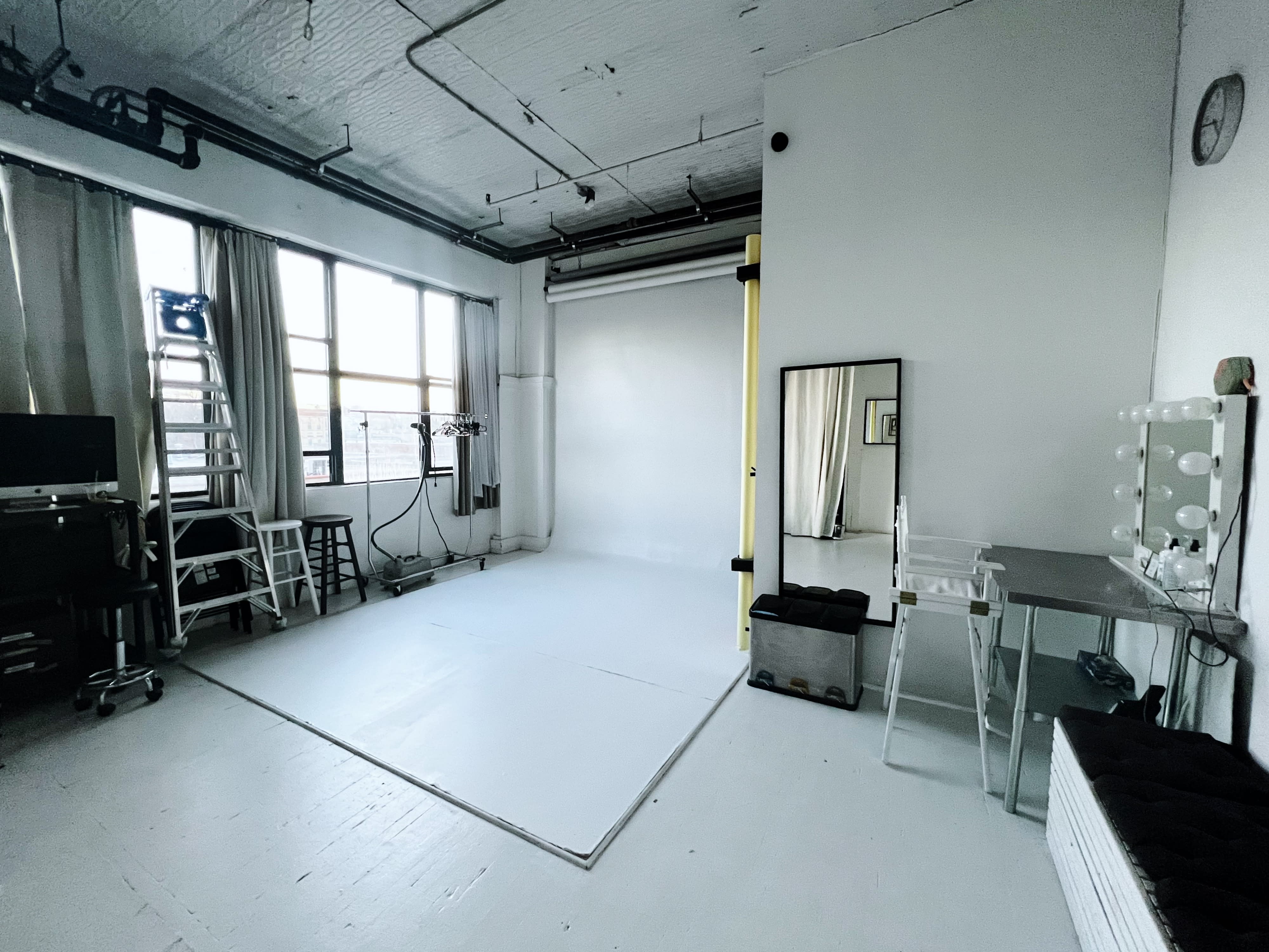 A photography studio features a backdrop with a ladder, various chairs, and soft natural light filtering through large windows.