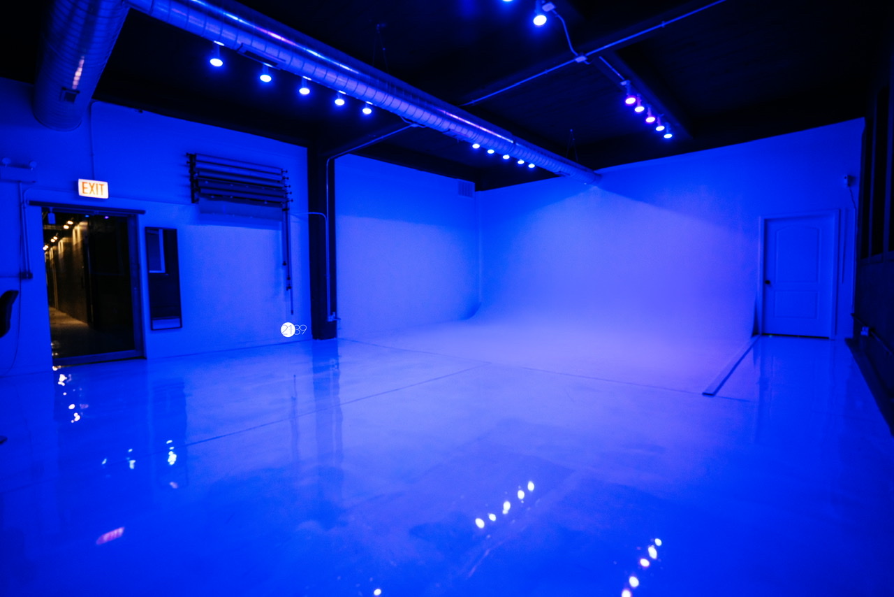 A video shoot takes place in a green-lit studio, featuring two individuals, one sitting and the other kneeling as they set up the scene.