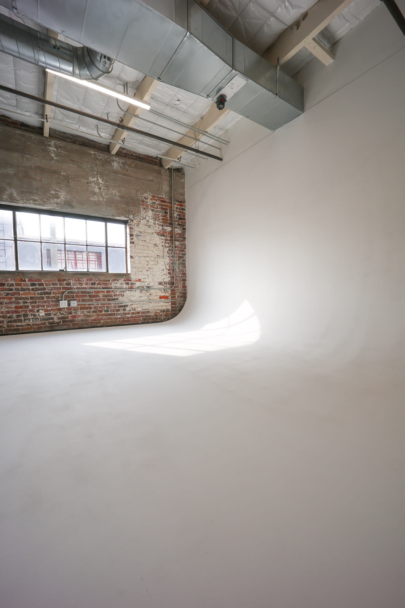 A man poses thoughtfully in a minimalist studio setting, standing next to a clear plastic chair while looking down at the ground.