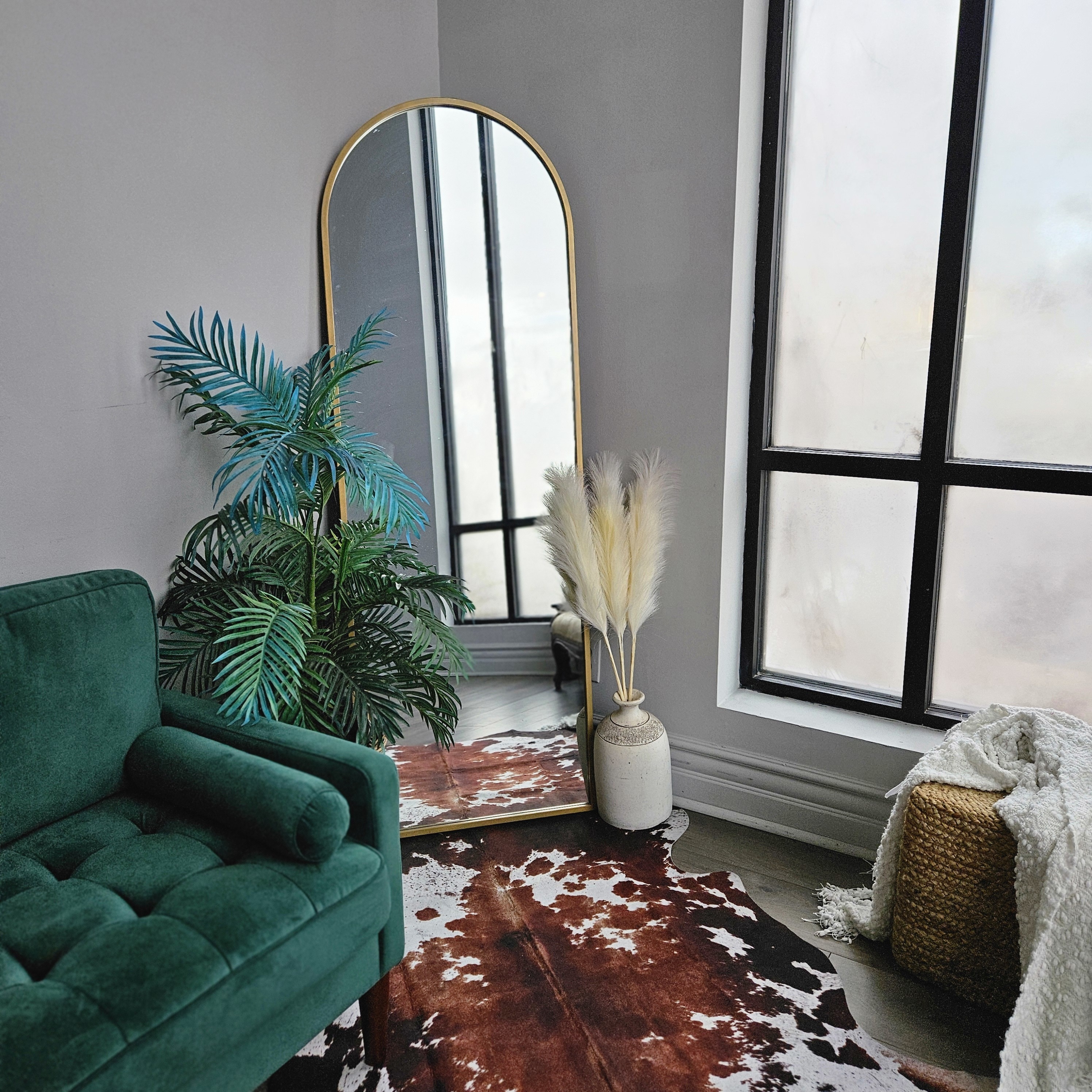 A woman sits cross-legged on a rug with her arms raised in a bright room featuring tall windows and plants.