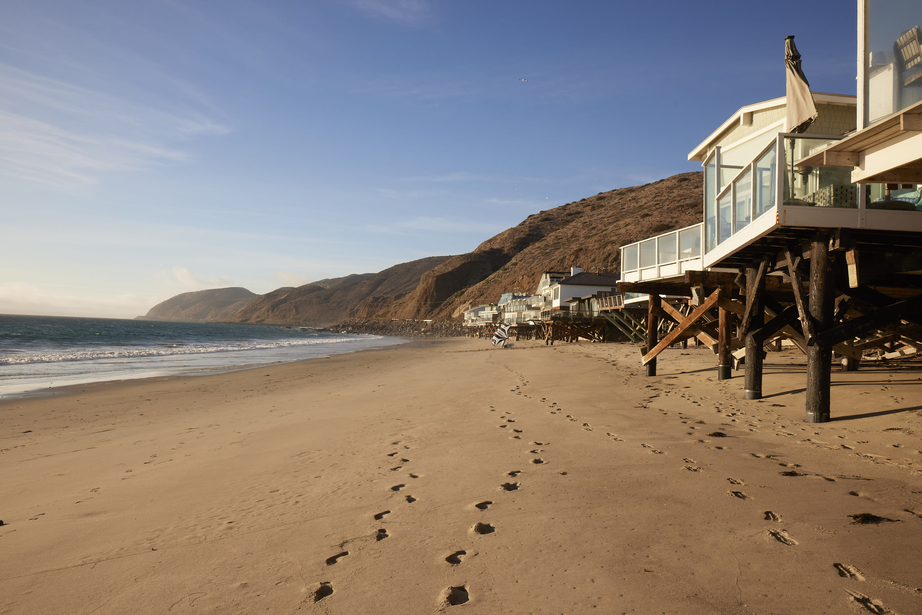 A photography shoot takes place on a wooden deck outside a triangular-roofed cabin overlooking the ocean at sunset.