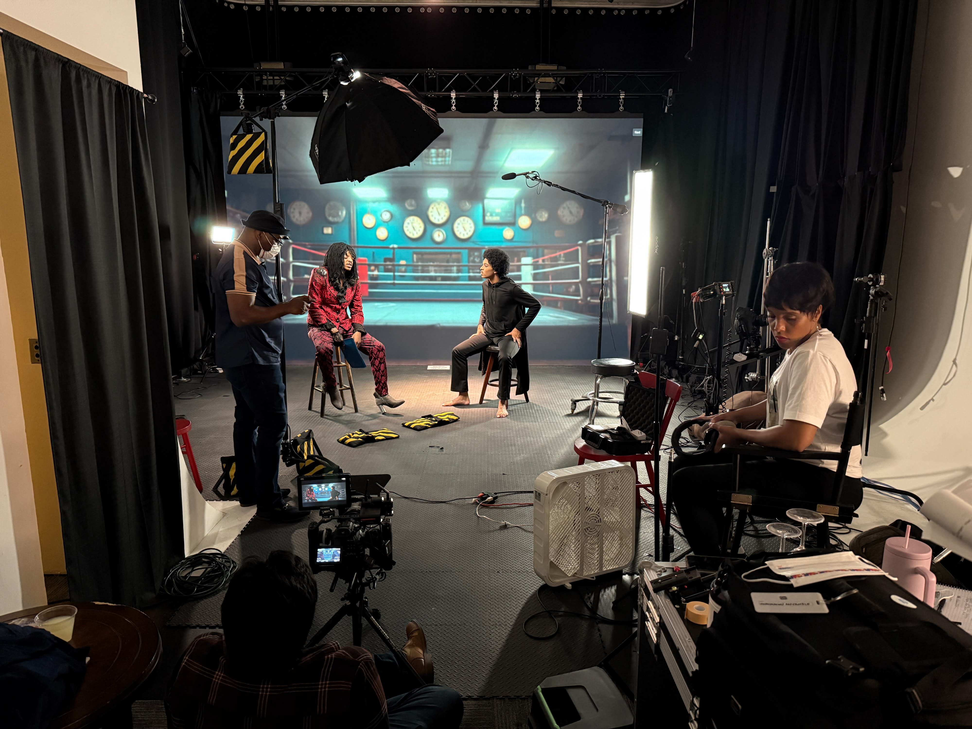 Two women pose in front of a camera while a crew member adjusts lighting in a studio setting.