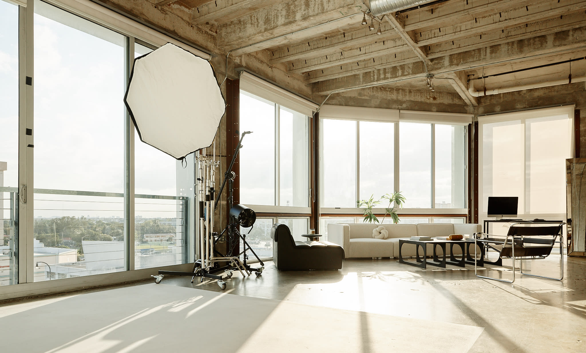 A model poses on one knee in a spacious studio with a blank backdrop and a large window overlooking the city.