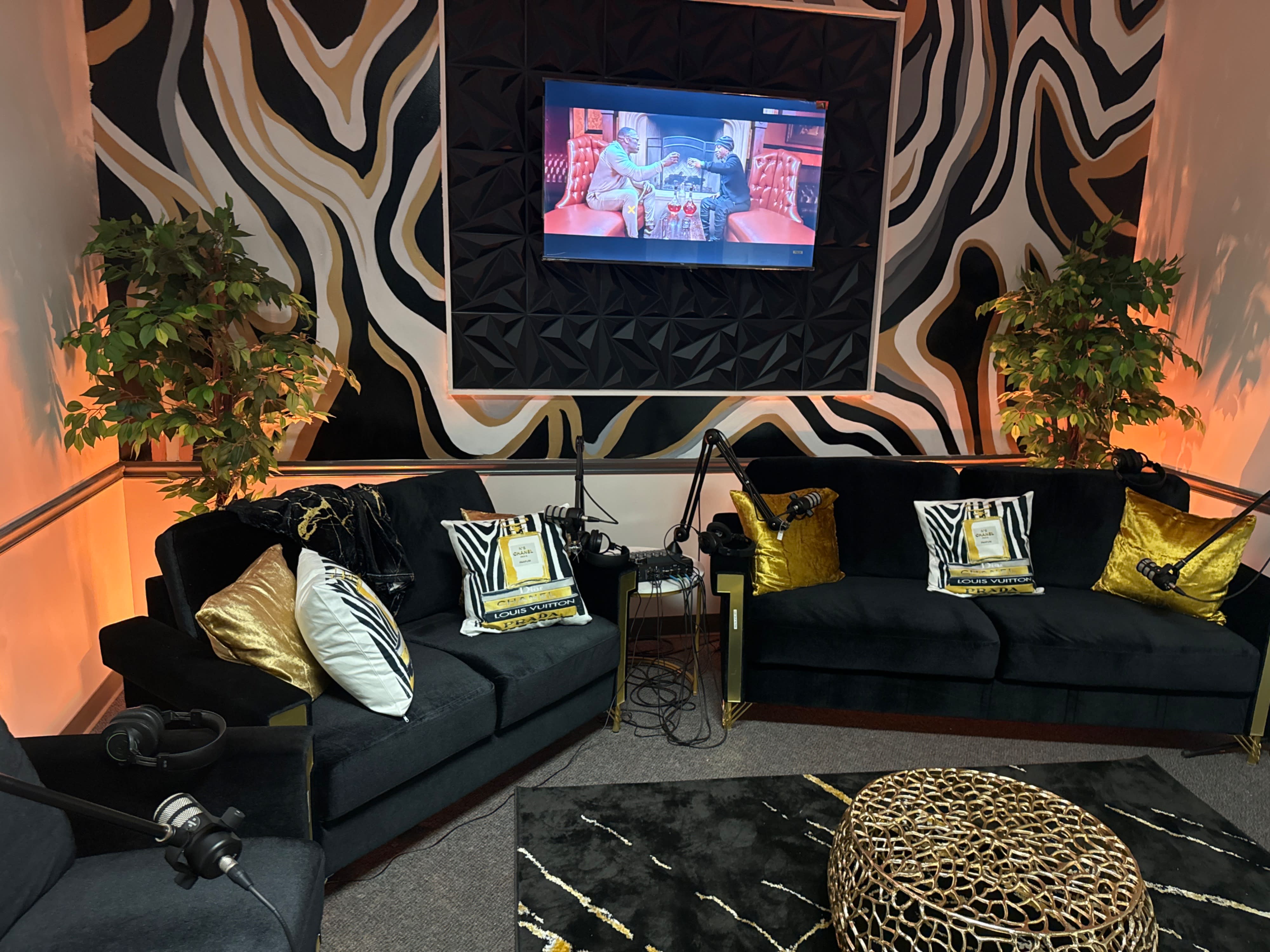 A podcast video shoot takes place with two hosts seated on a patterned couch, preparing for recording alongside microphones and lighting equipment.