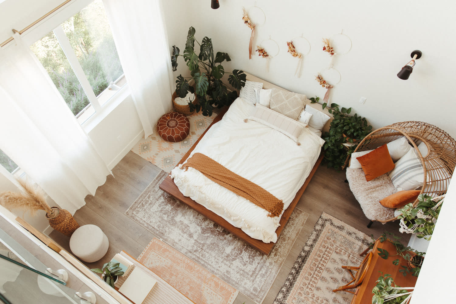 A woman sits on a neatly made bed in a brightly lit room, surrounded by decorative wall hangings and indoor plants.