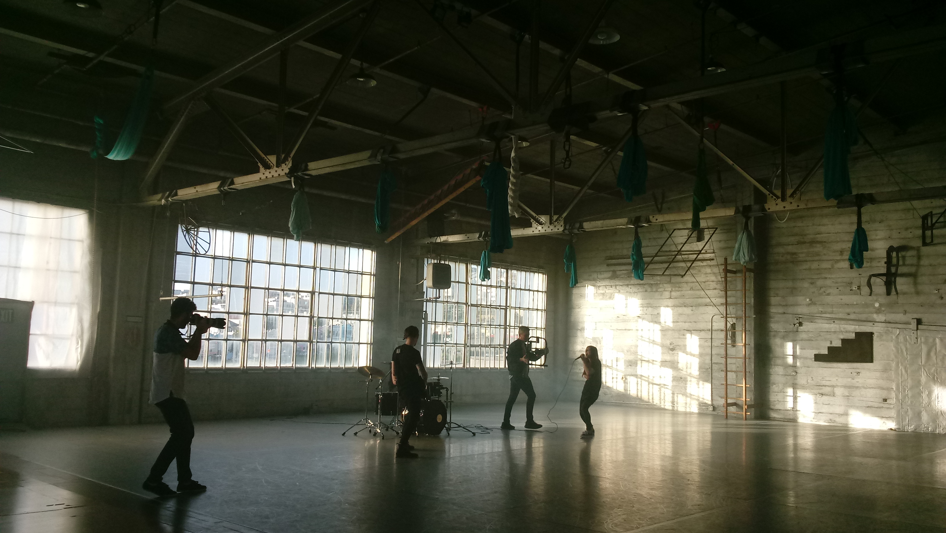 A video shoot takes place in a spacious studio, with a crew filming a person seated on a chair, illuminated by natural light streaming through large windows.