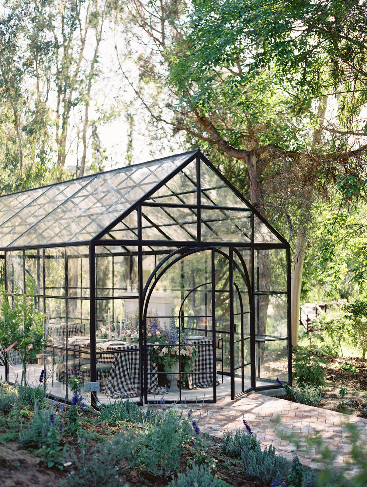 A couple stands together, smiling and toasting with glasses in a beautifully decorated greenhouse set up for a rehearsal dinner.
