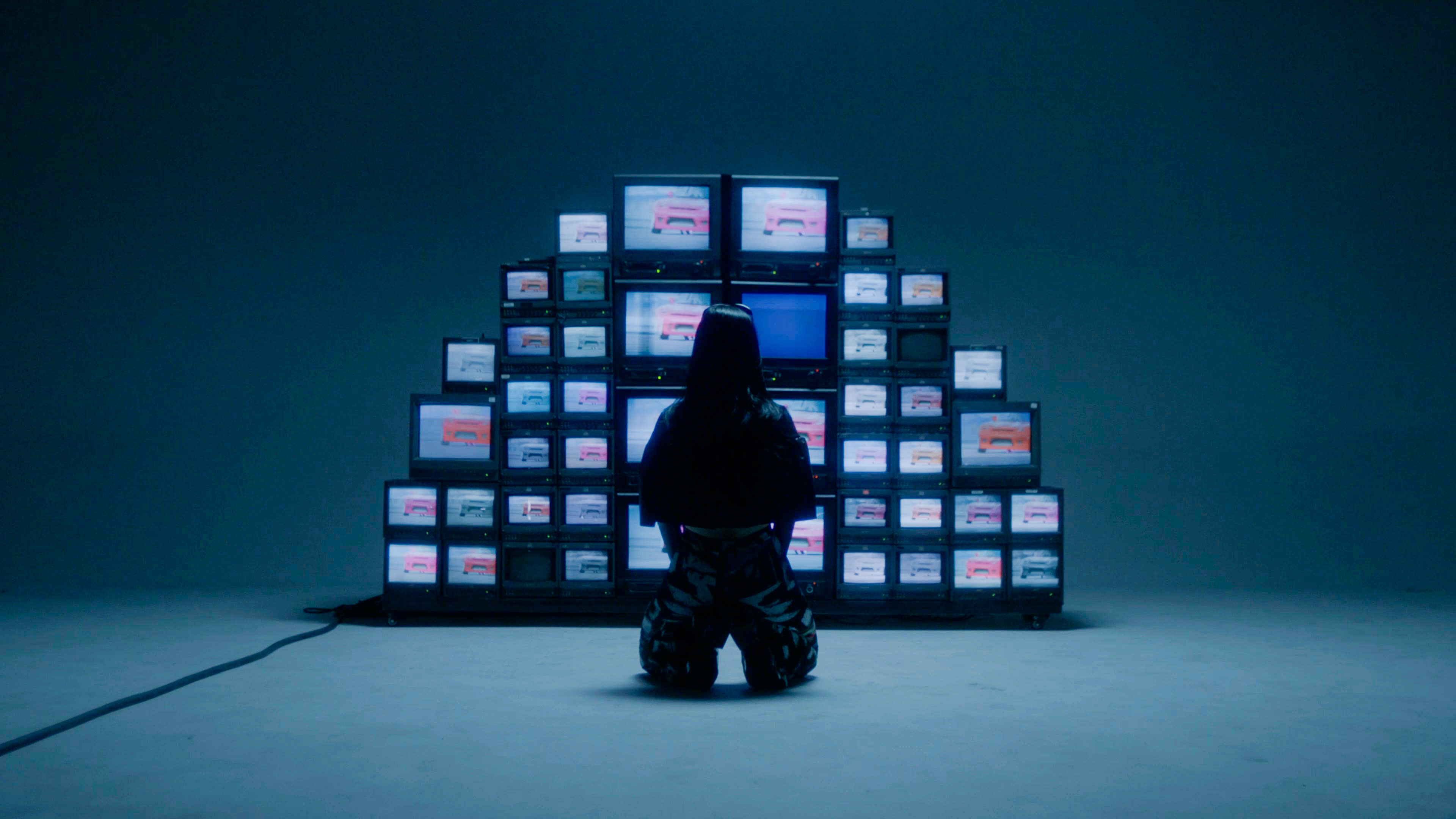 A person stands in front of a wall of vintage televisions while another individual operates a camera on a tripod.