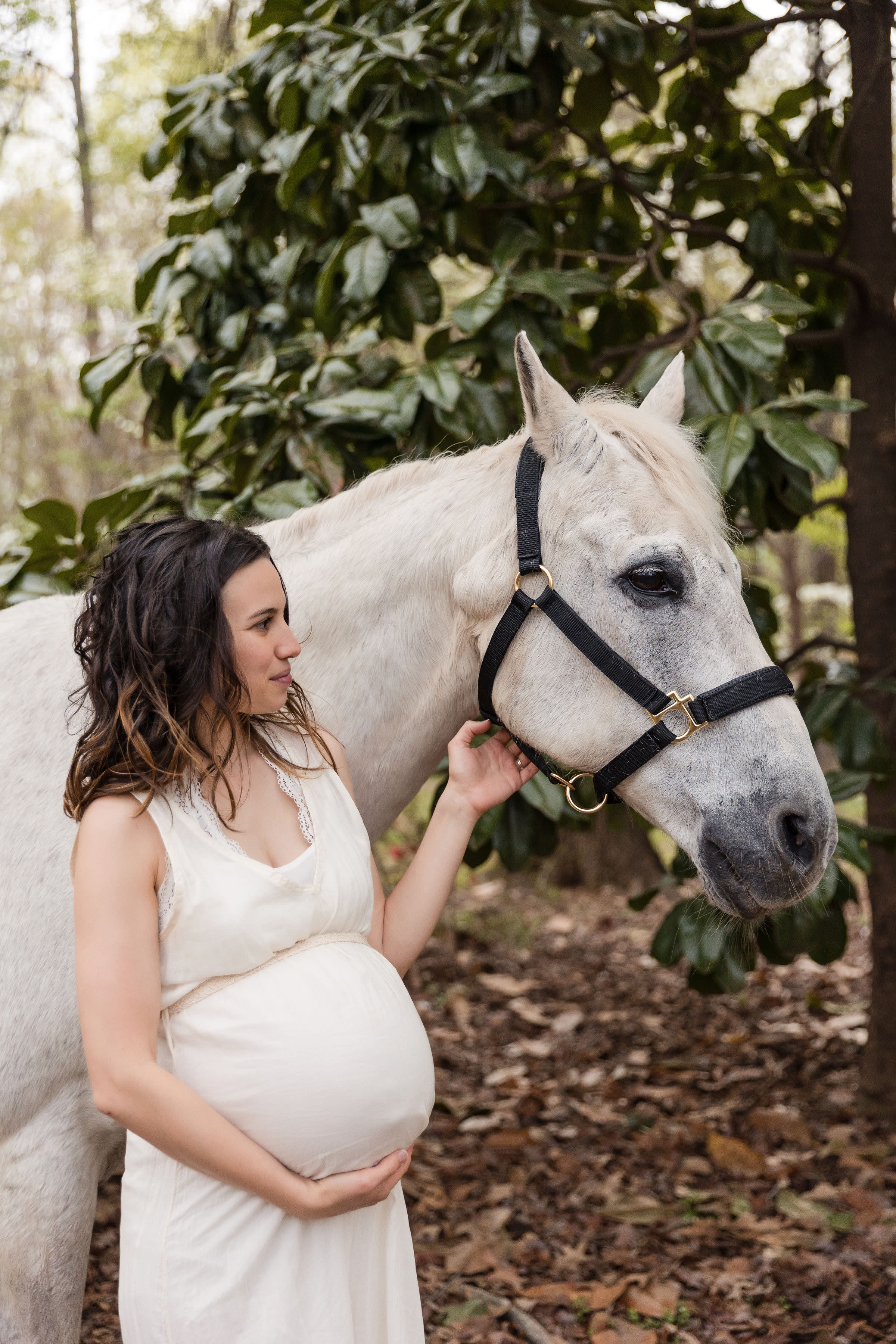 A pregnant woman kneels in a grassy area beside a white horse under the trees.