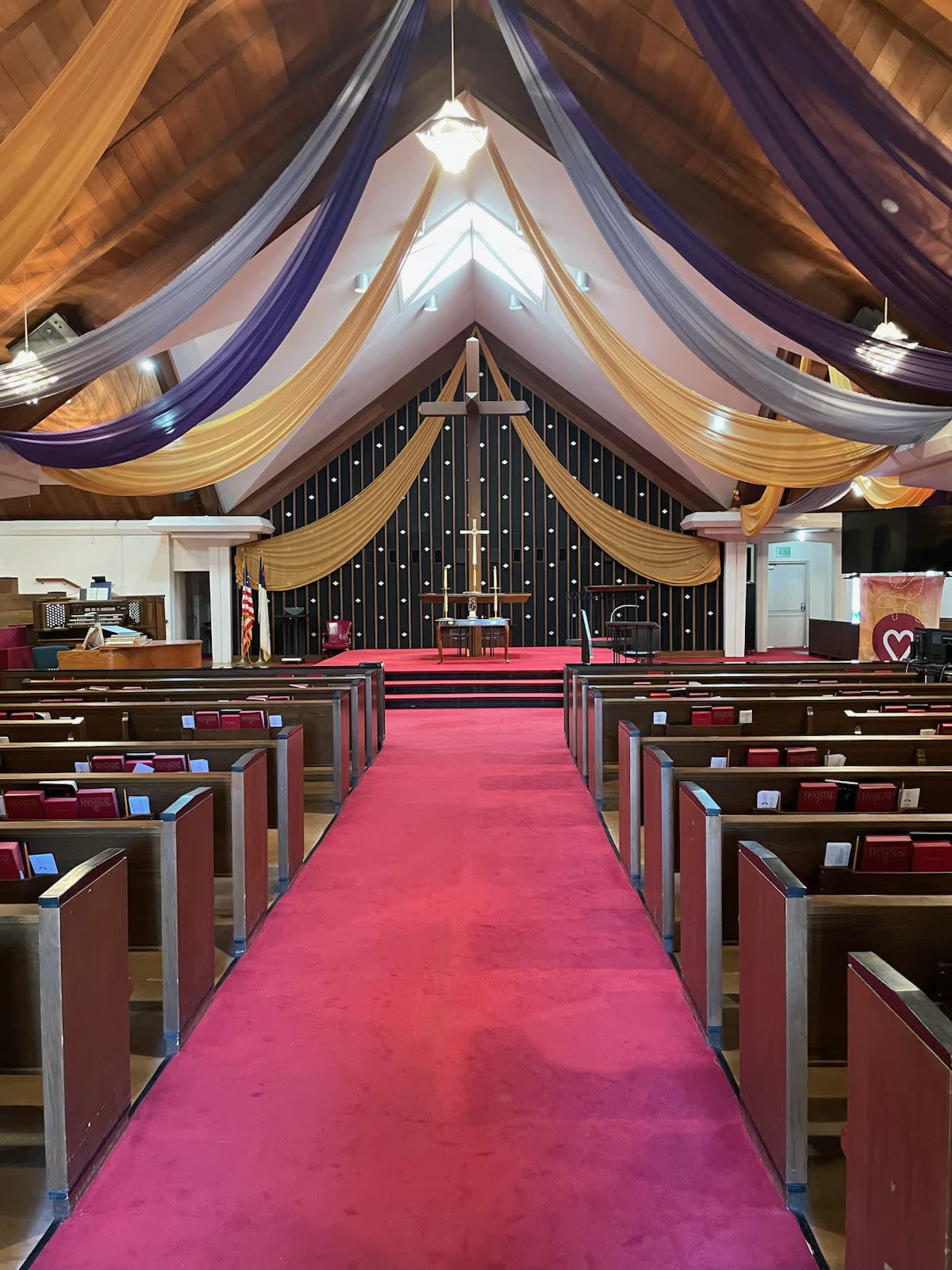 A couple exchanges vows at a wedding ceremony under a large wooden cross in a church adorned with floral arrangements.