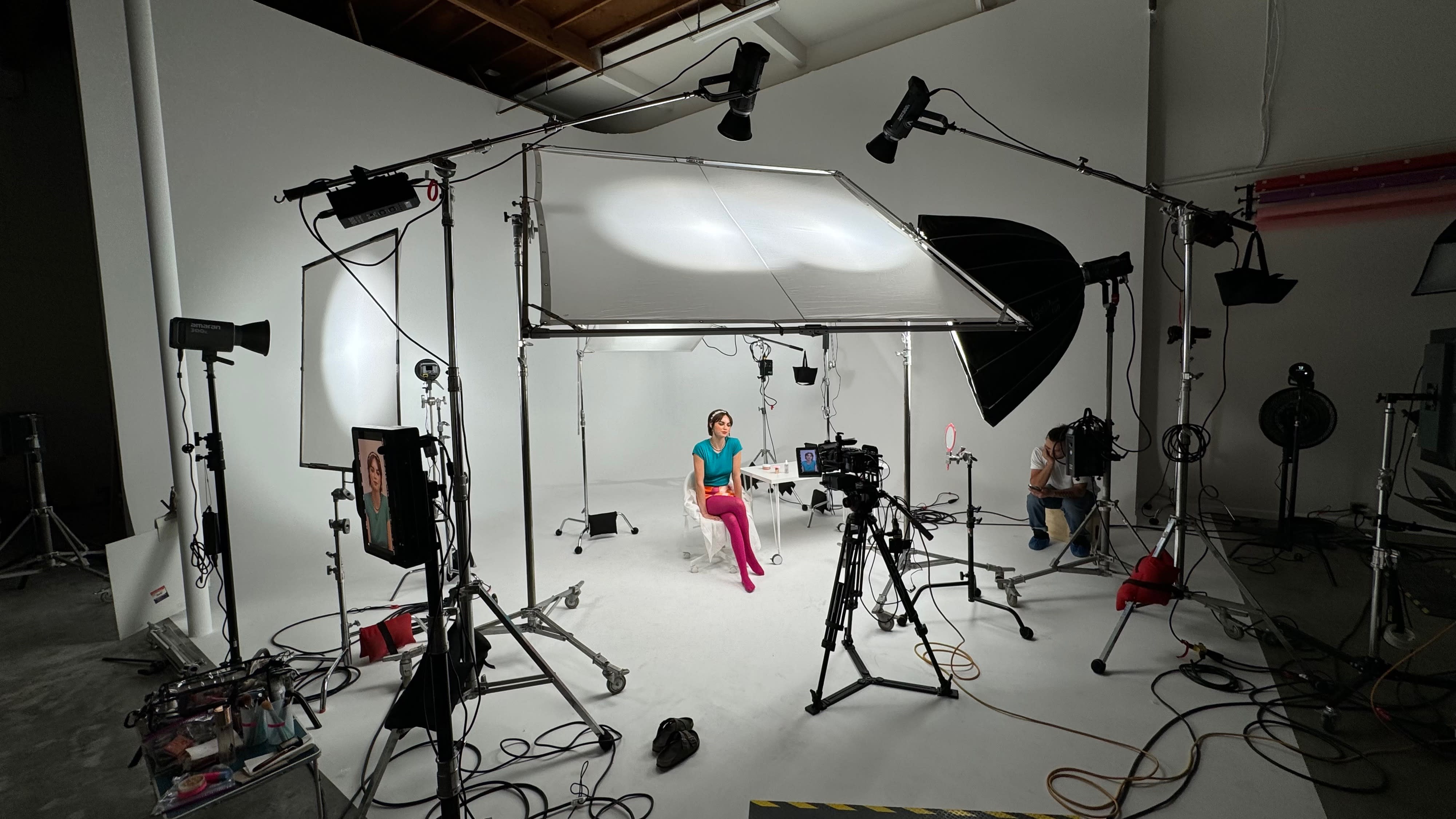 Two individuals are seated at a table in front of a white backdrop, while a third person operates a camera and a fourth holds a light source during a video shoot.