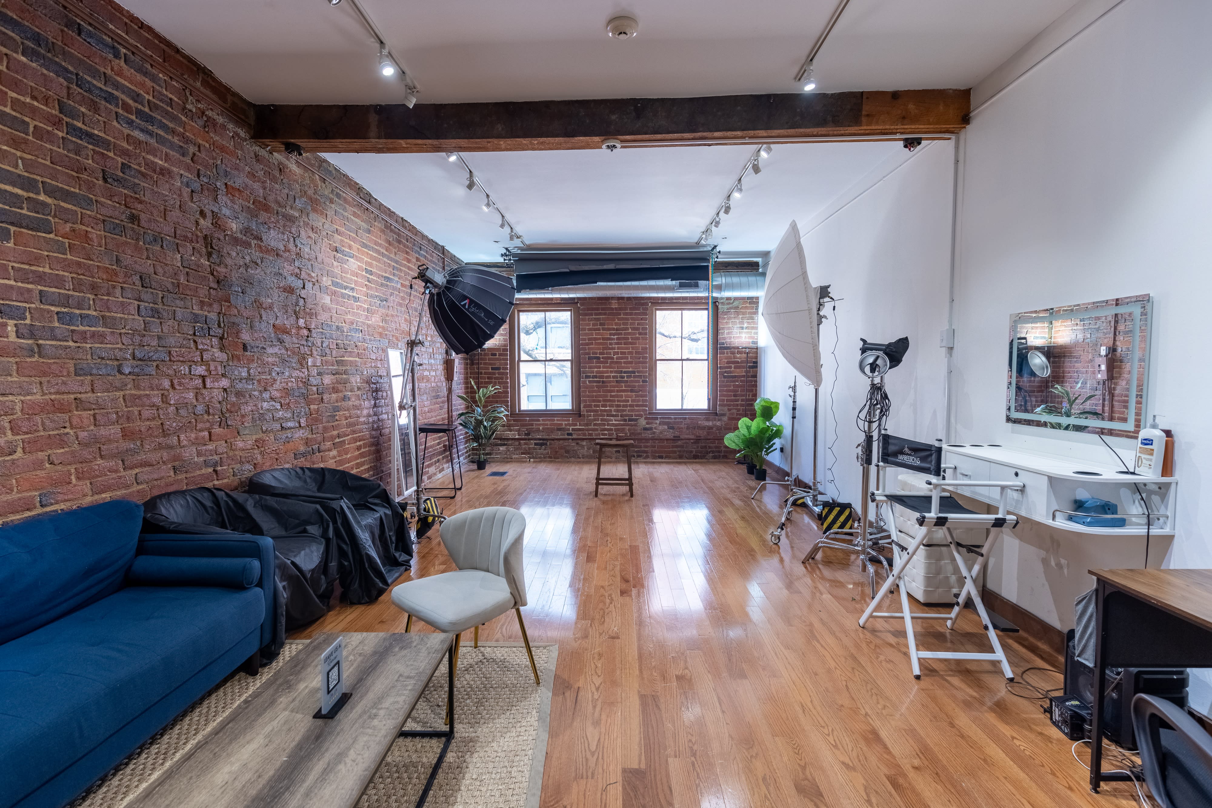 Four black stools are arranged in a row on a wooden floor under natural light from large windows in a warehouse-like space with brick walls.