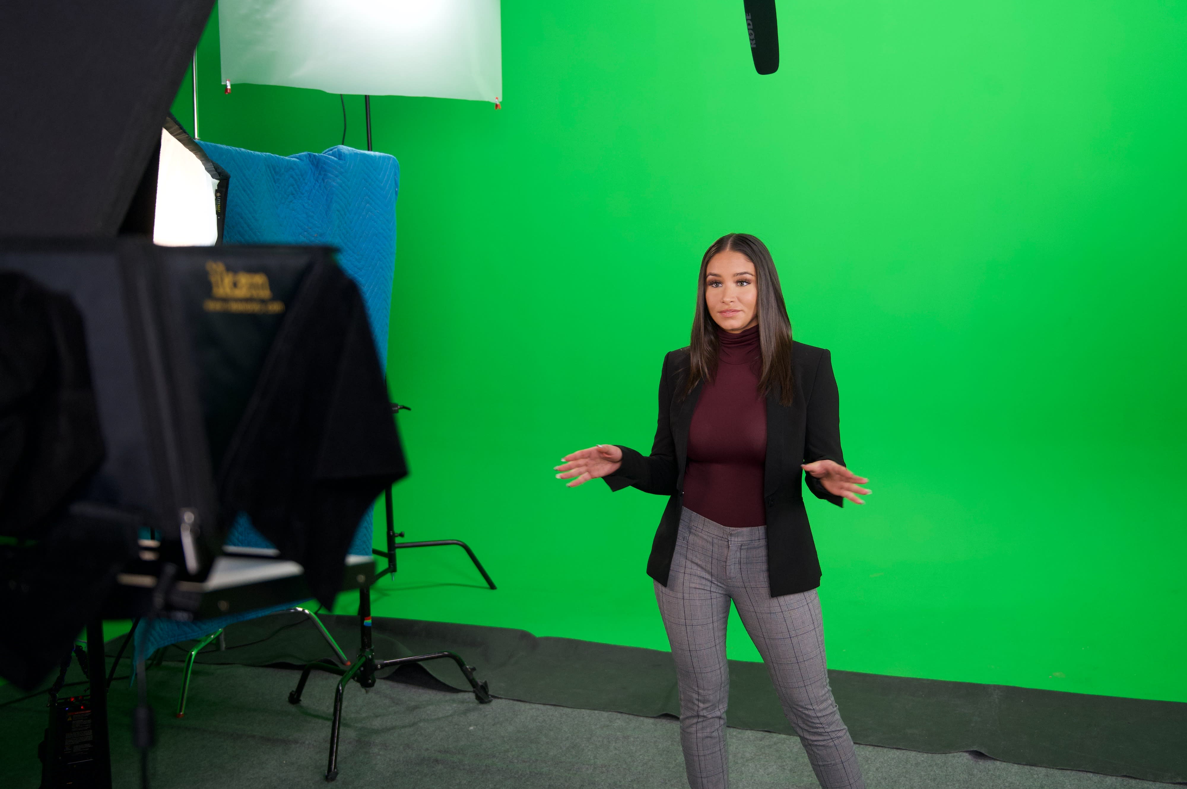 A person sits on a brown chair in a green screen studio while being filmed by a camera set up in front of them.
