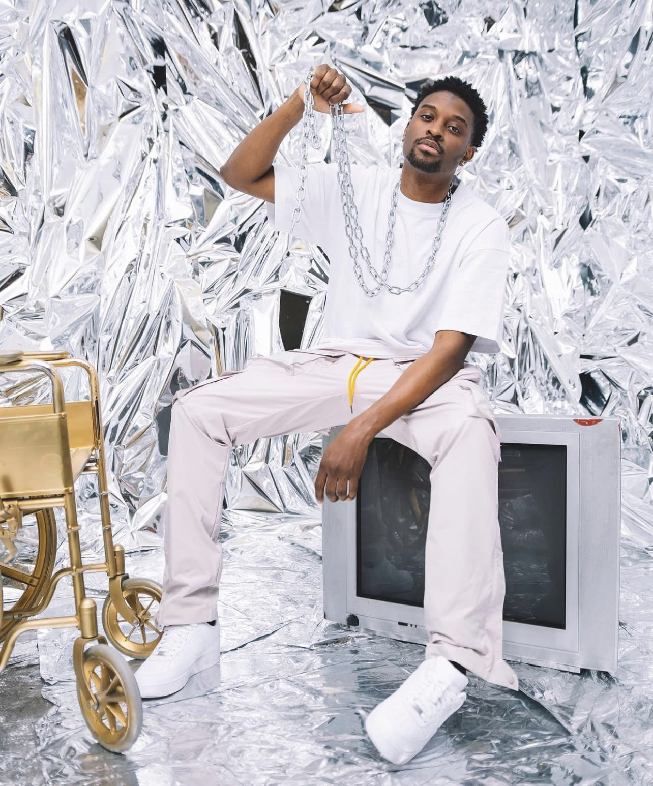 A person dressed in a shiny silver outfit sits on a television while holding a boombox against a backdrop of crinkled metallic material.