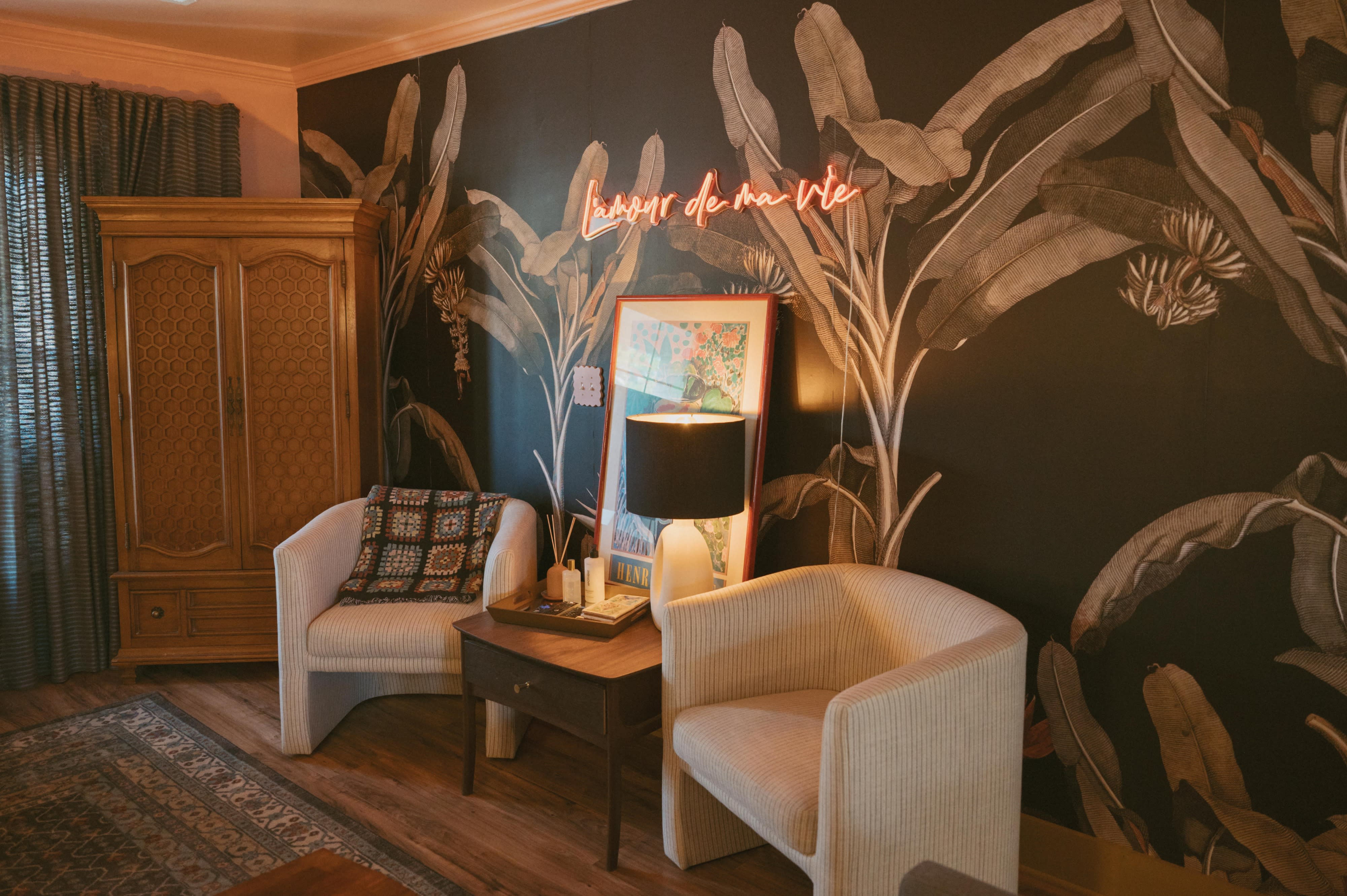 A woman sits thoughtfully on a couch in a stylish room decorated with plants and a neon sign.