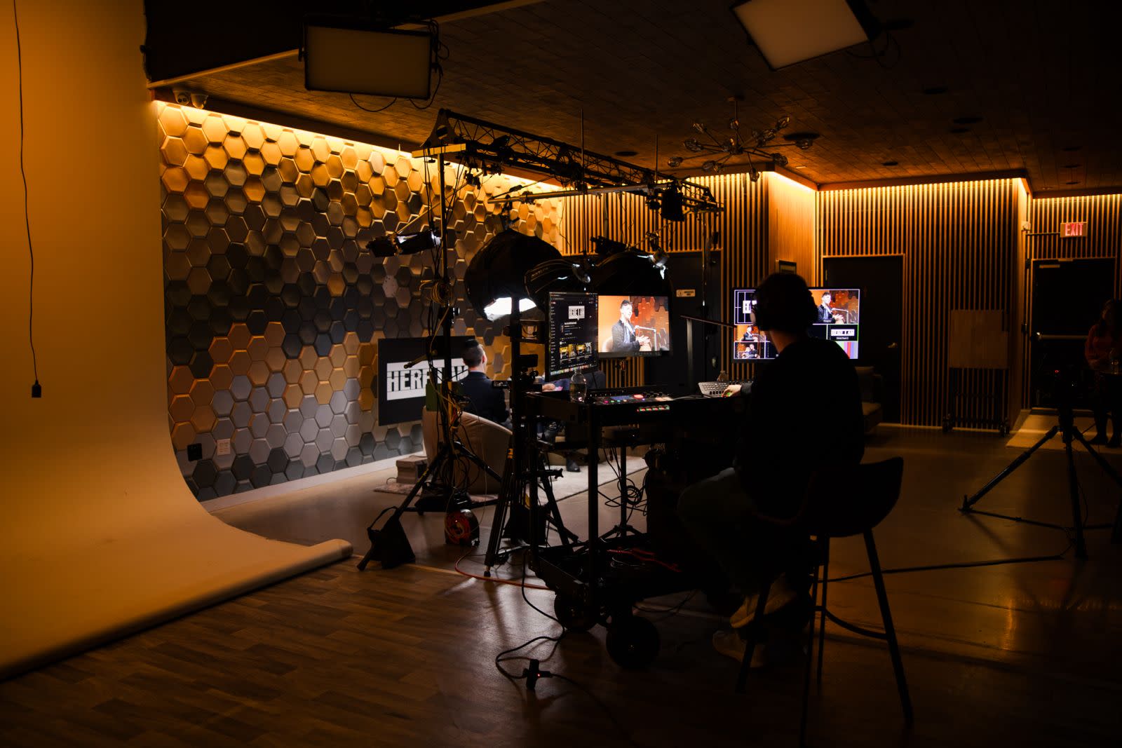 A video production crew sets up equipment in a blank studio while a subject prepares for filming.