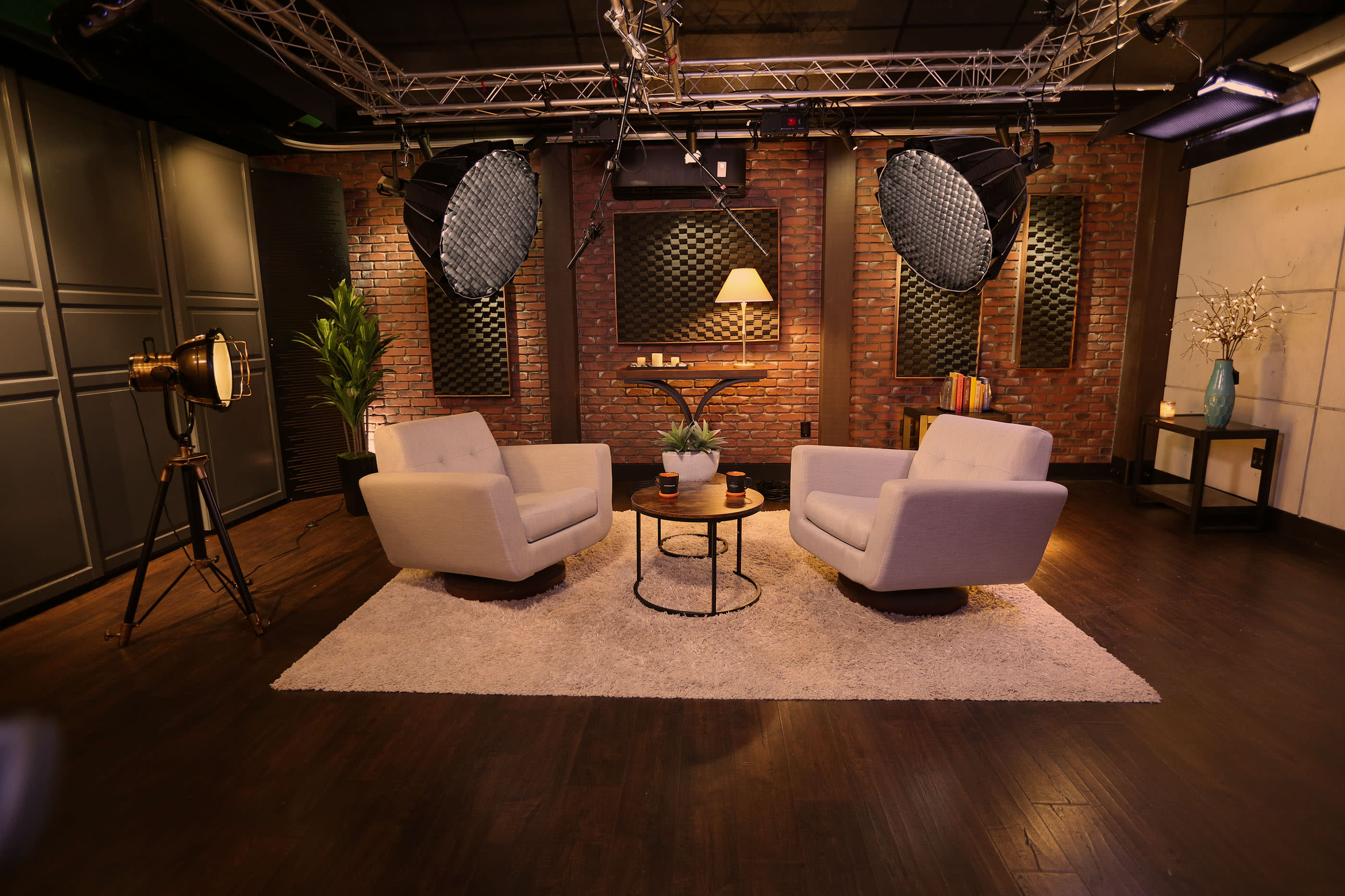 Two men sit on a couch during a video interview, with a camera displaying their conversation and promotional materials in the background.