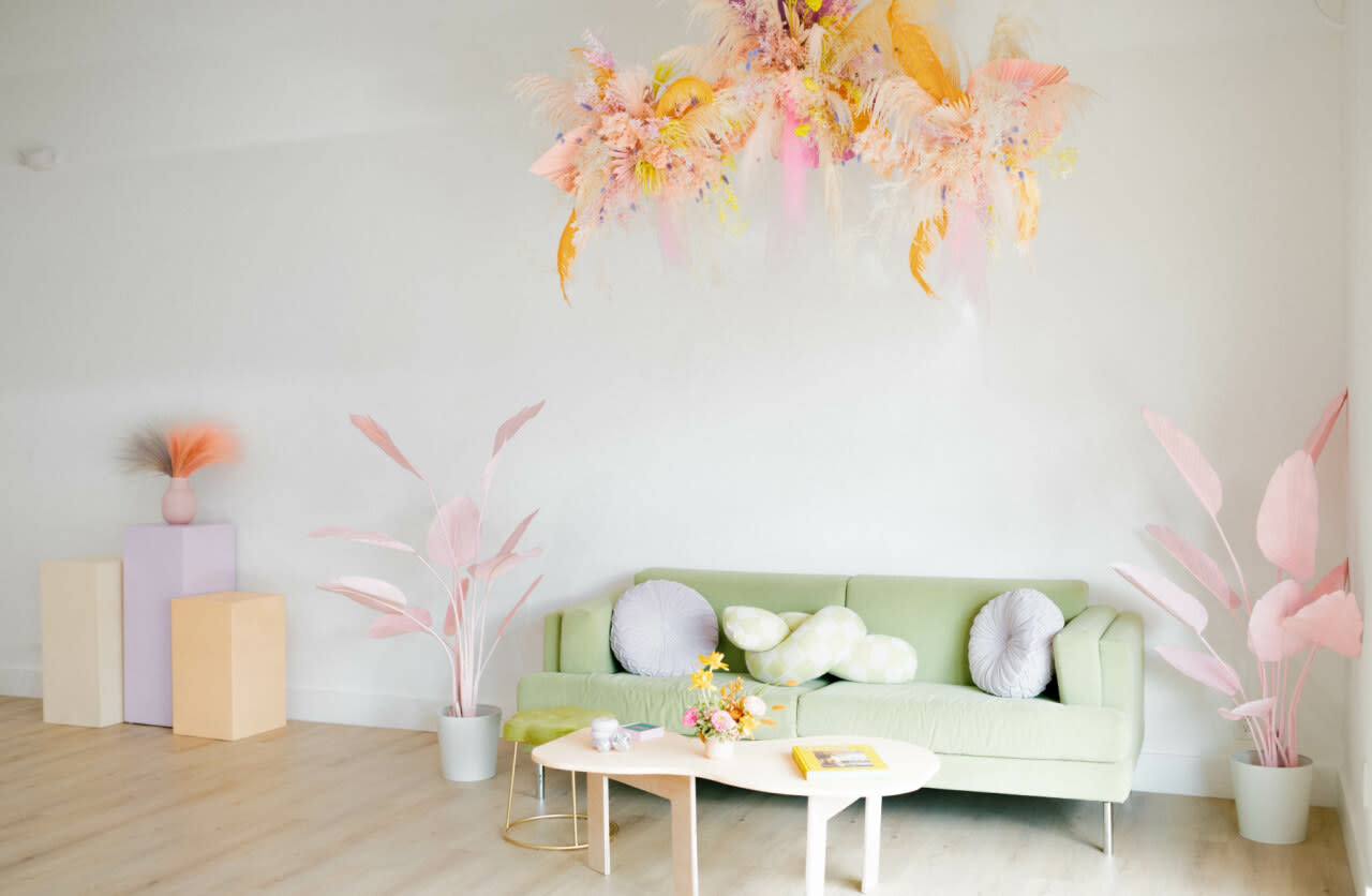 The scene features a decorated room for a baby shower, showcasing a vibrant floral arrangement above a green sofa and a coffee table with a magazine.
