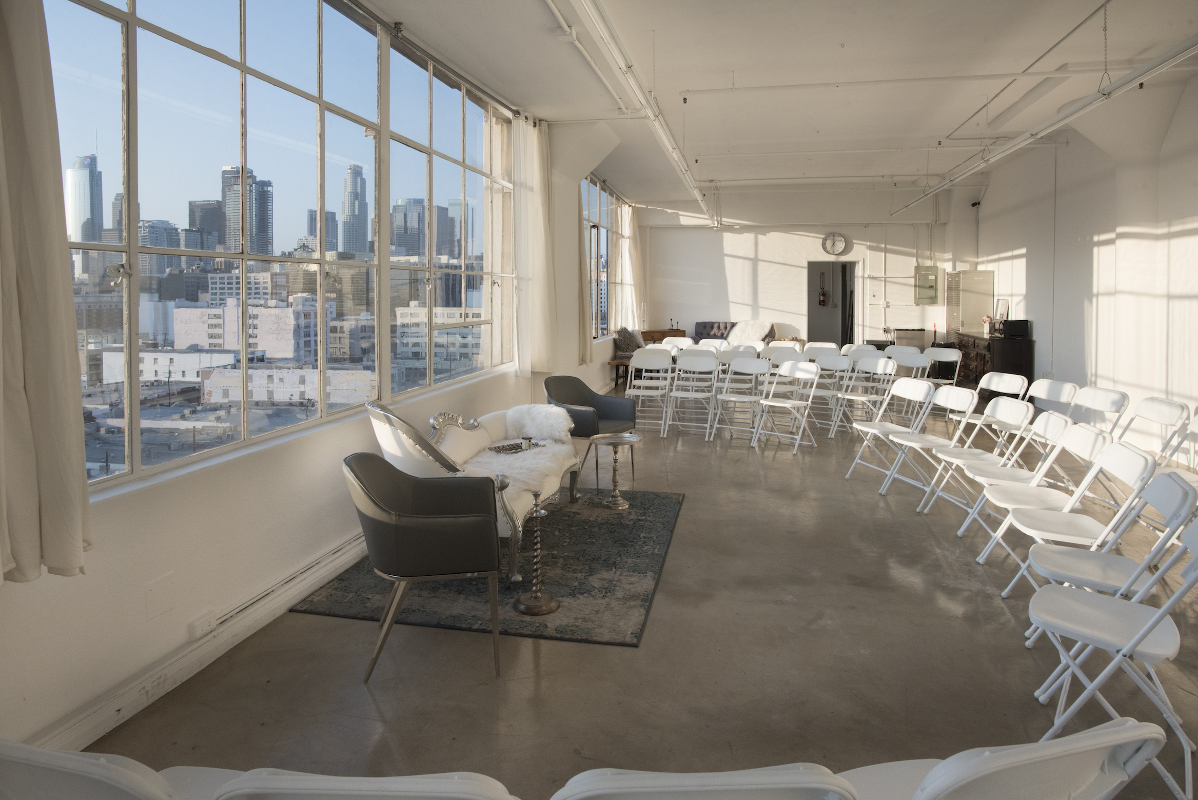 A modern photo shoot setup features a white couch and decorative plants in an industrial space with large windows overlooking a city skyline.