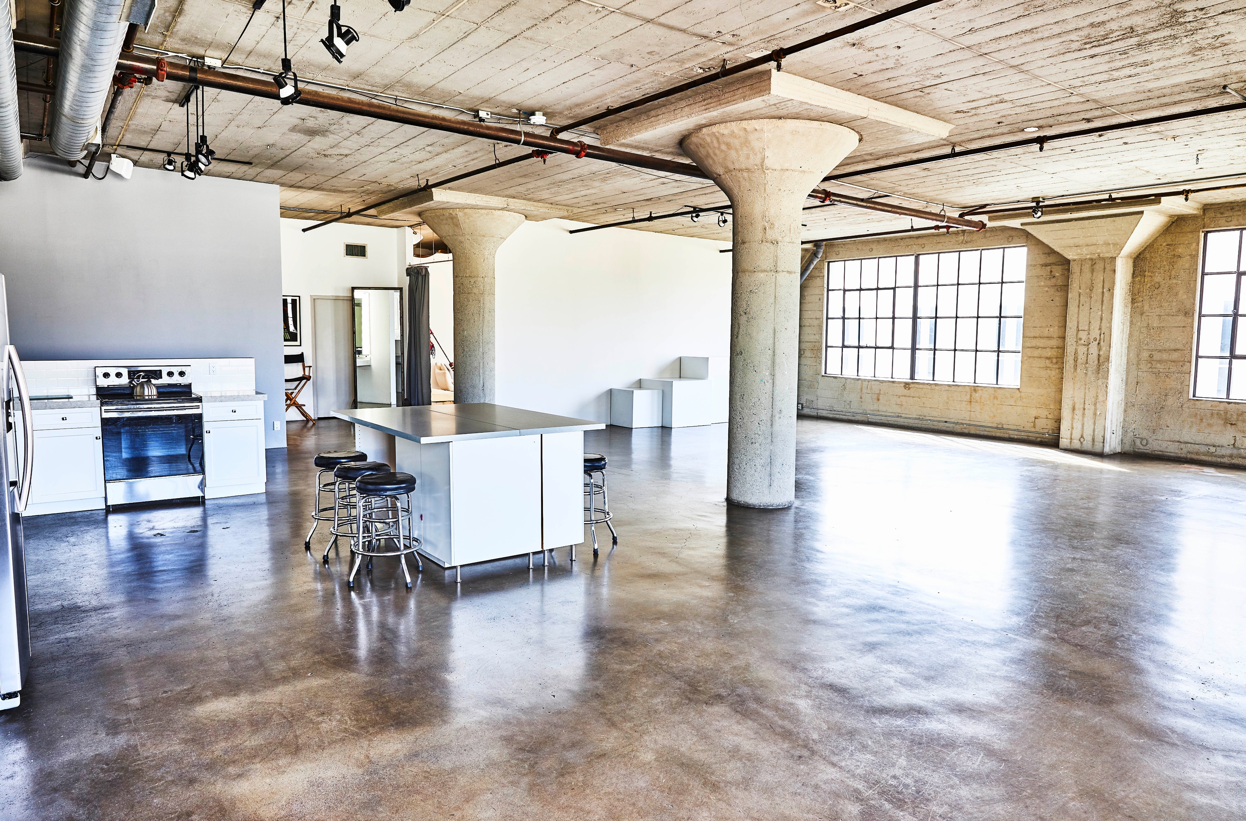 A photoshoot captures two models posing casually in a spacious industrial room, with one reclining in a chair and the other seated beside her.