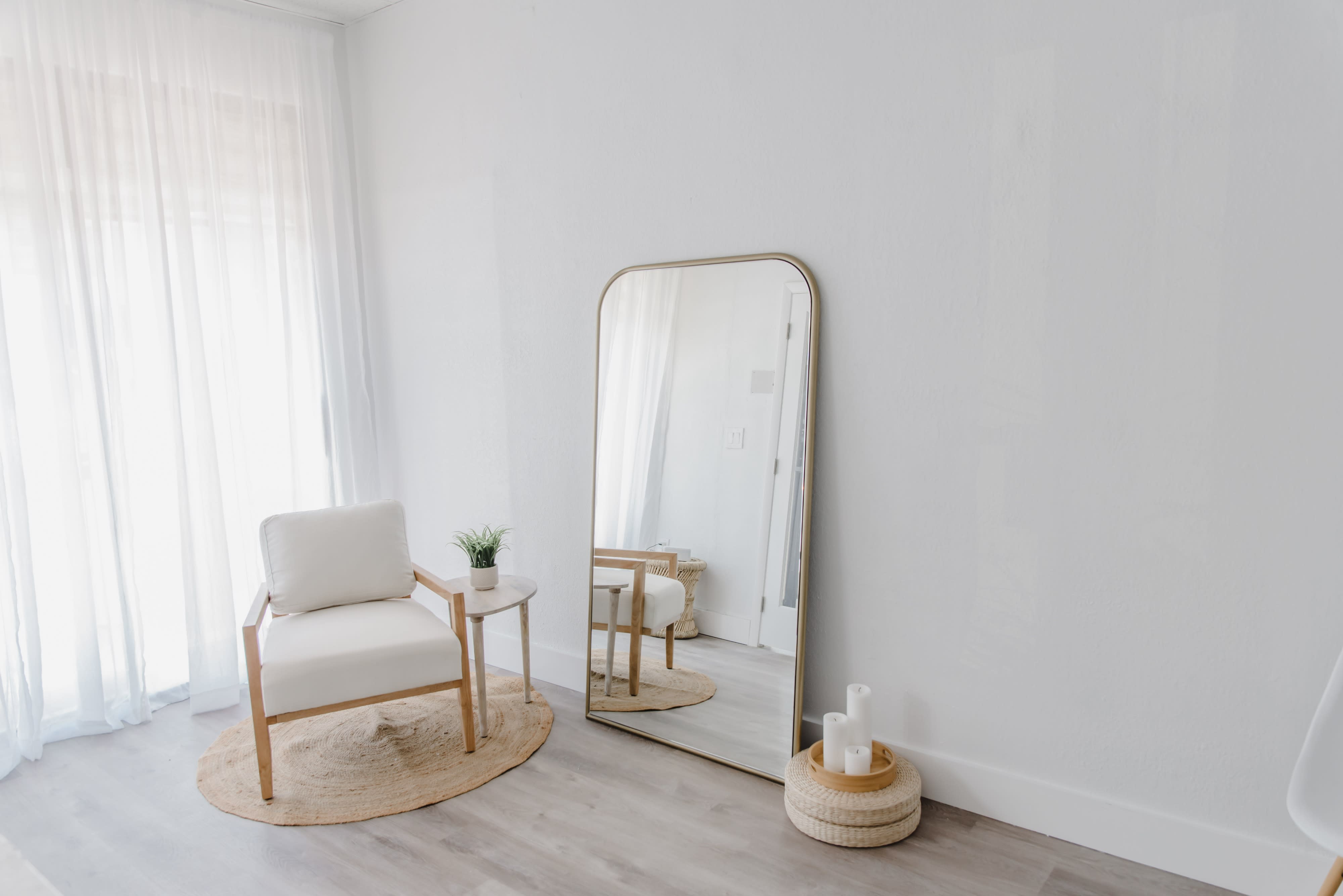 A person takes a selfie in a mirror while standing in a light-filled room with neutral decor.